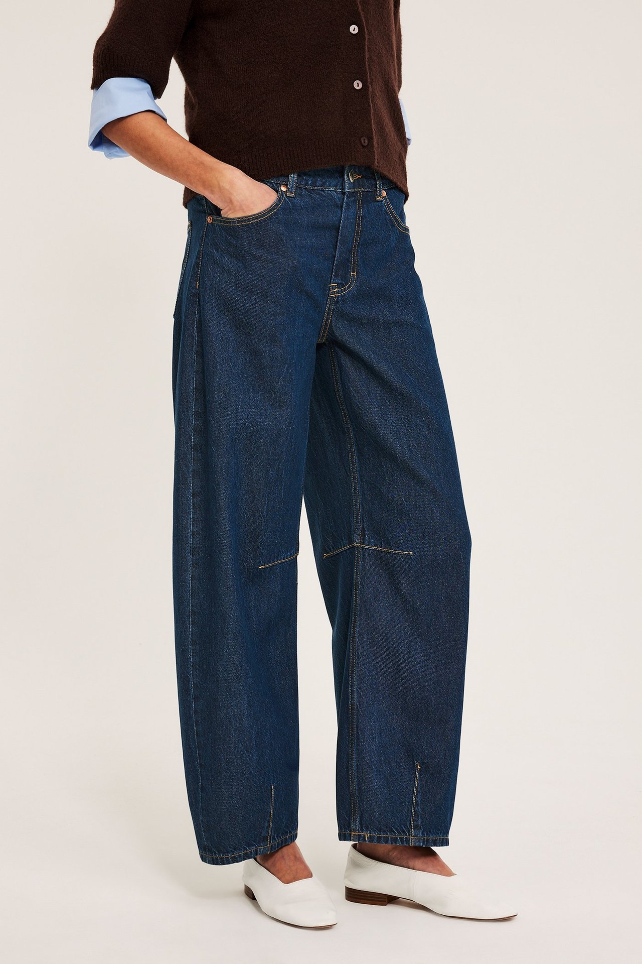 Frontvy av kvinna i mörkblå barrel fit vida jeans, ljusblå skjorta, brun kofta, vita platta skor.