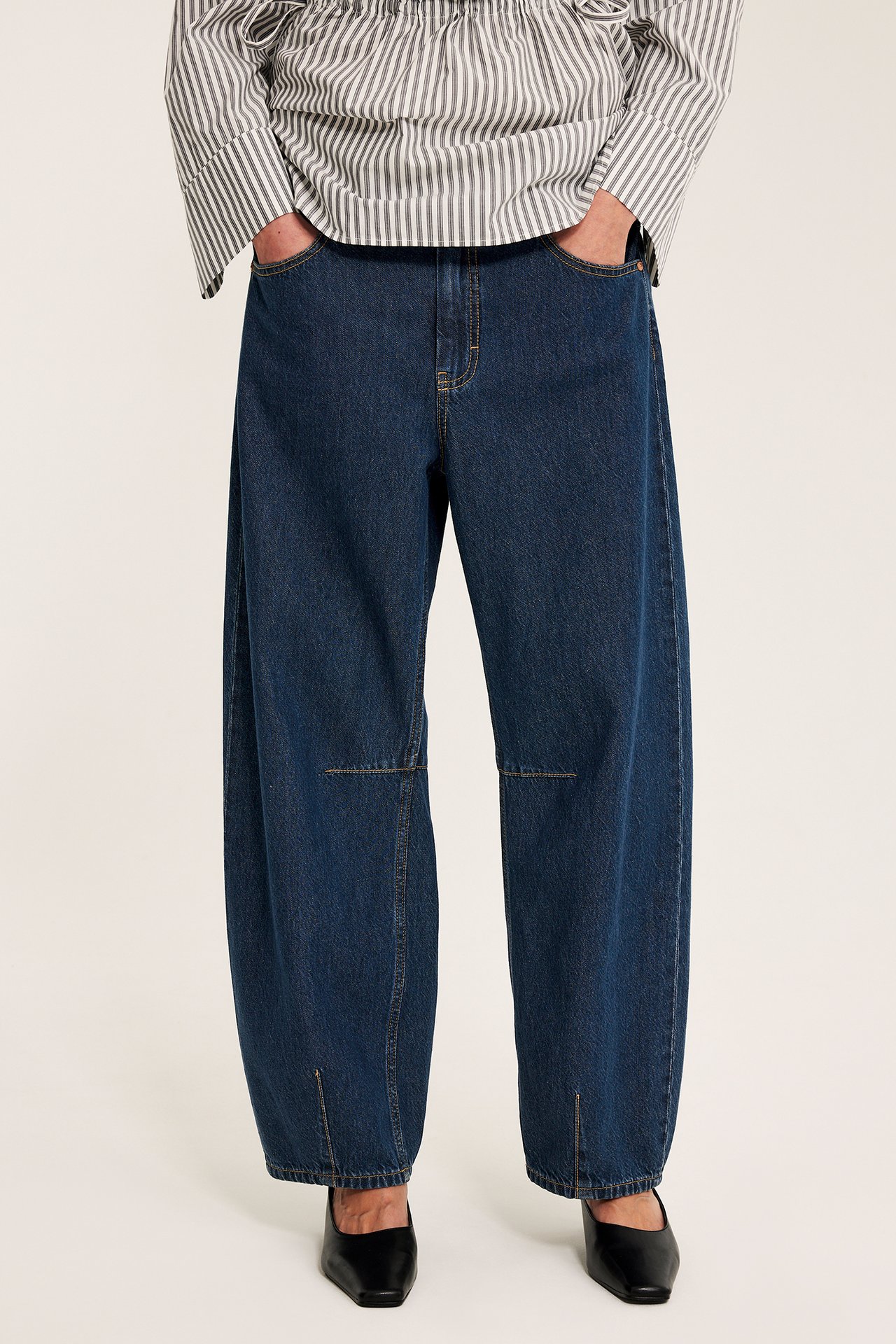 Forfra, kvinne i mørkeblå barrel-fit vide jeans med knesømmer og ankelslisser, stripete topp og svarte sko.