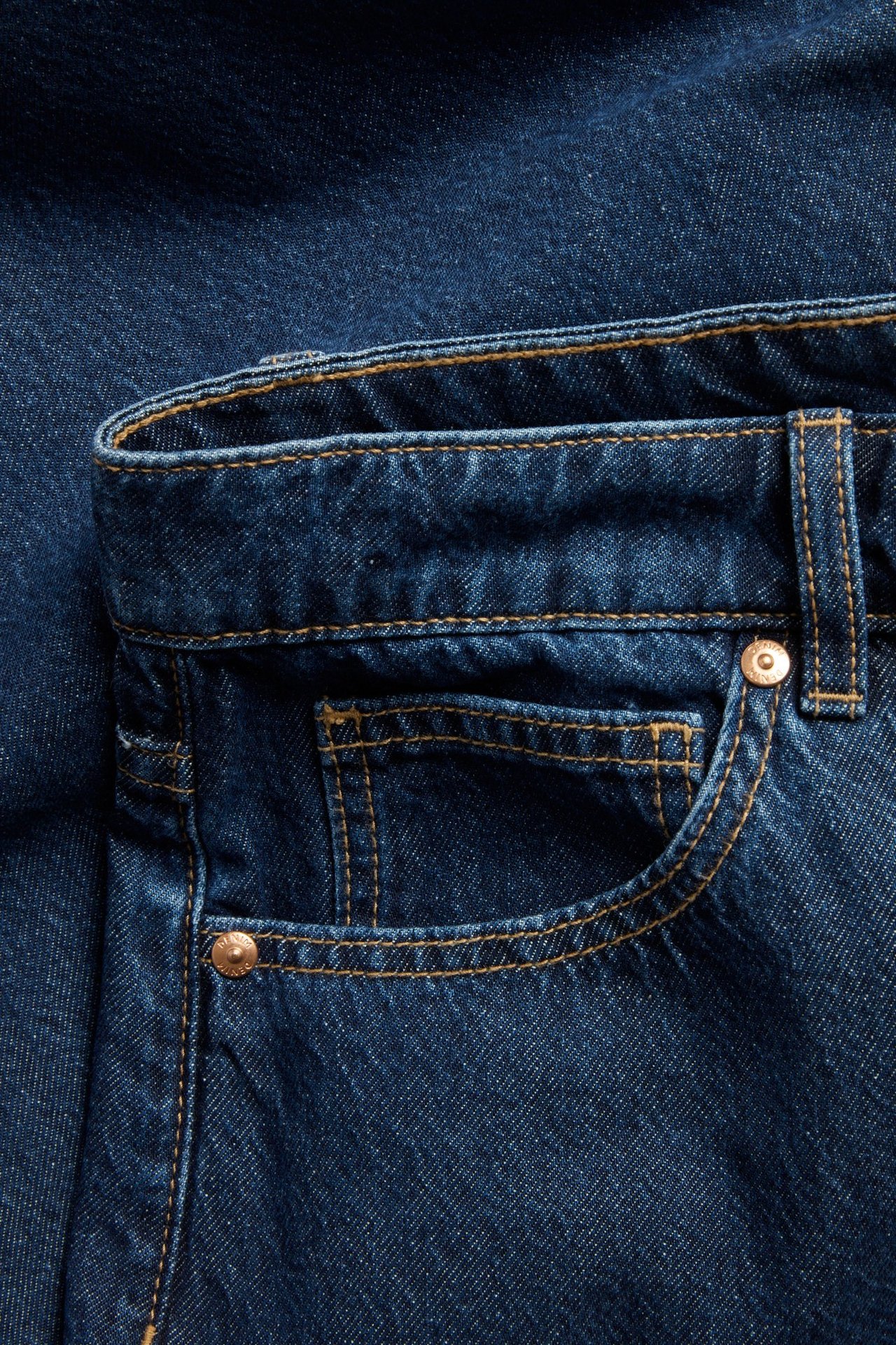 Zbliżenie na ciemnoniebieskie jeansy barrel fit z pomarańczowymi szwami i brązowymi nitami przy przedniej kieszeni.