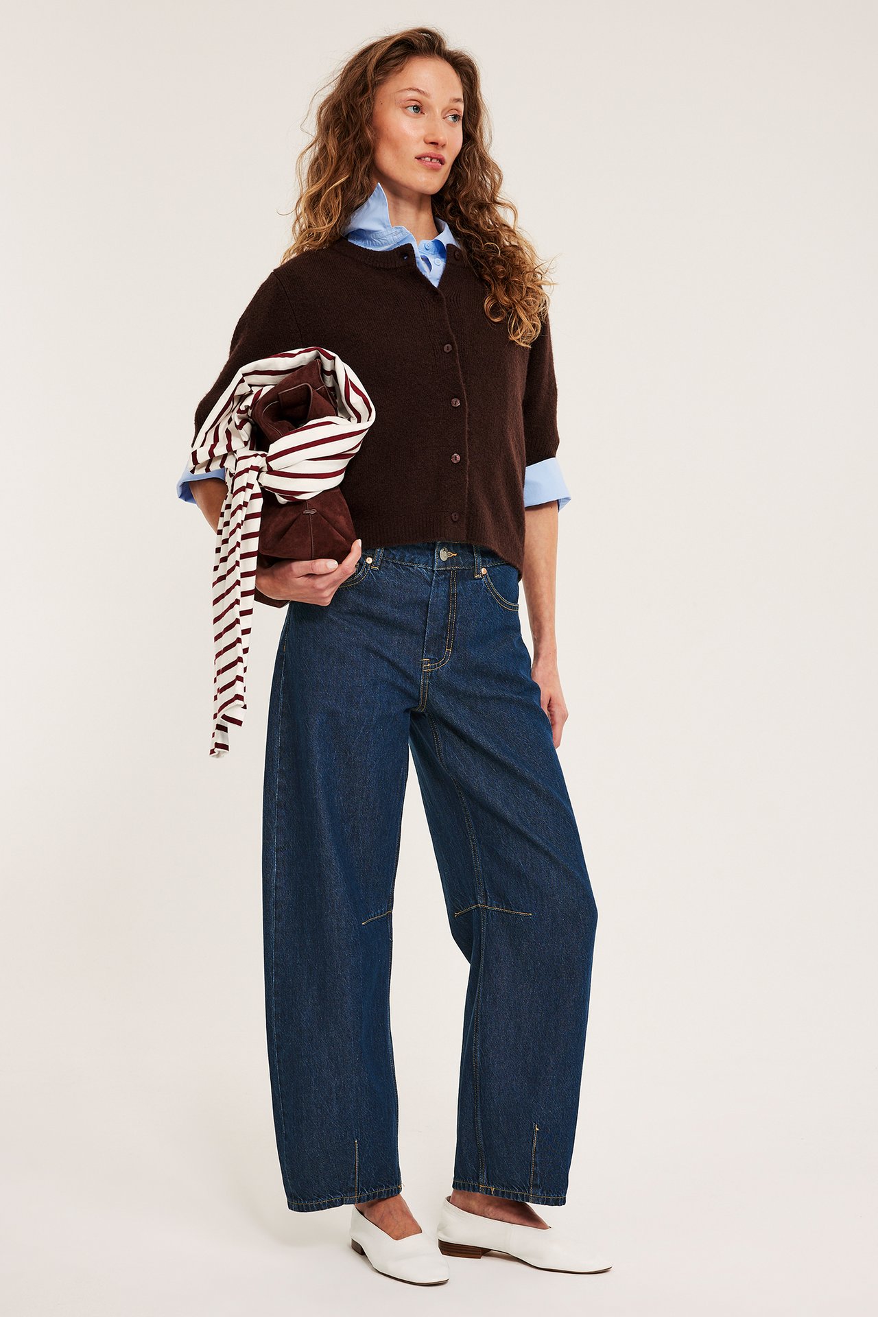 Nainen tummansinisissä barrel-farkuissa, ruskea neuletakki sinisen paidan päällä, pitelee ruskeaa laukkua, huivi.