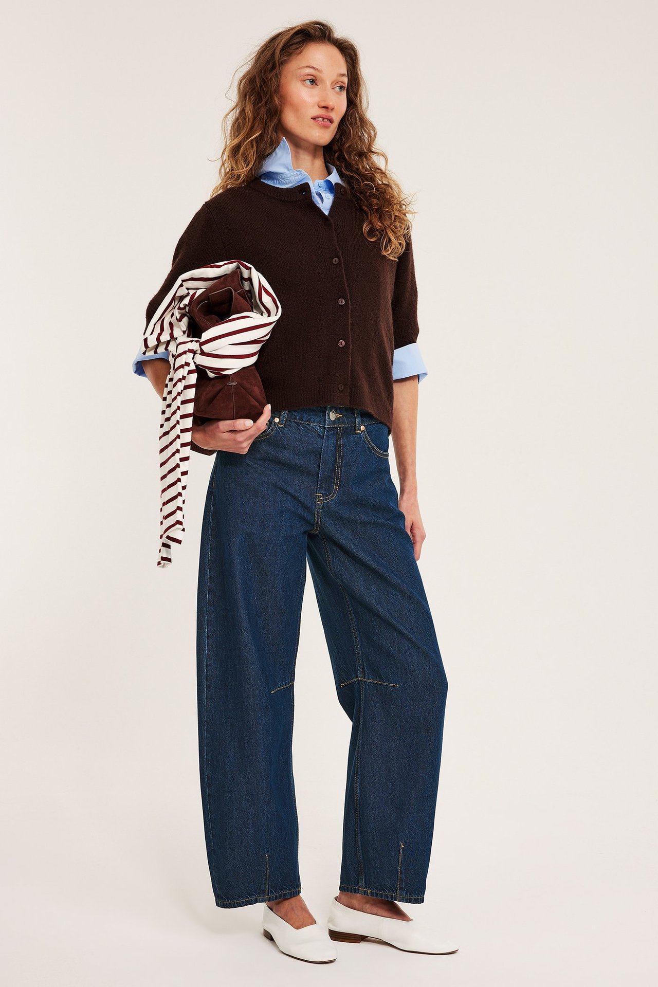 Kvinna i mörkblå barrel-jeans, brun kofta över blå skjorta, håller brun väska med sjal.