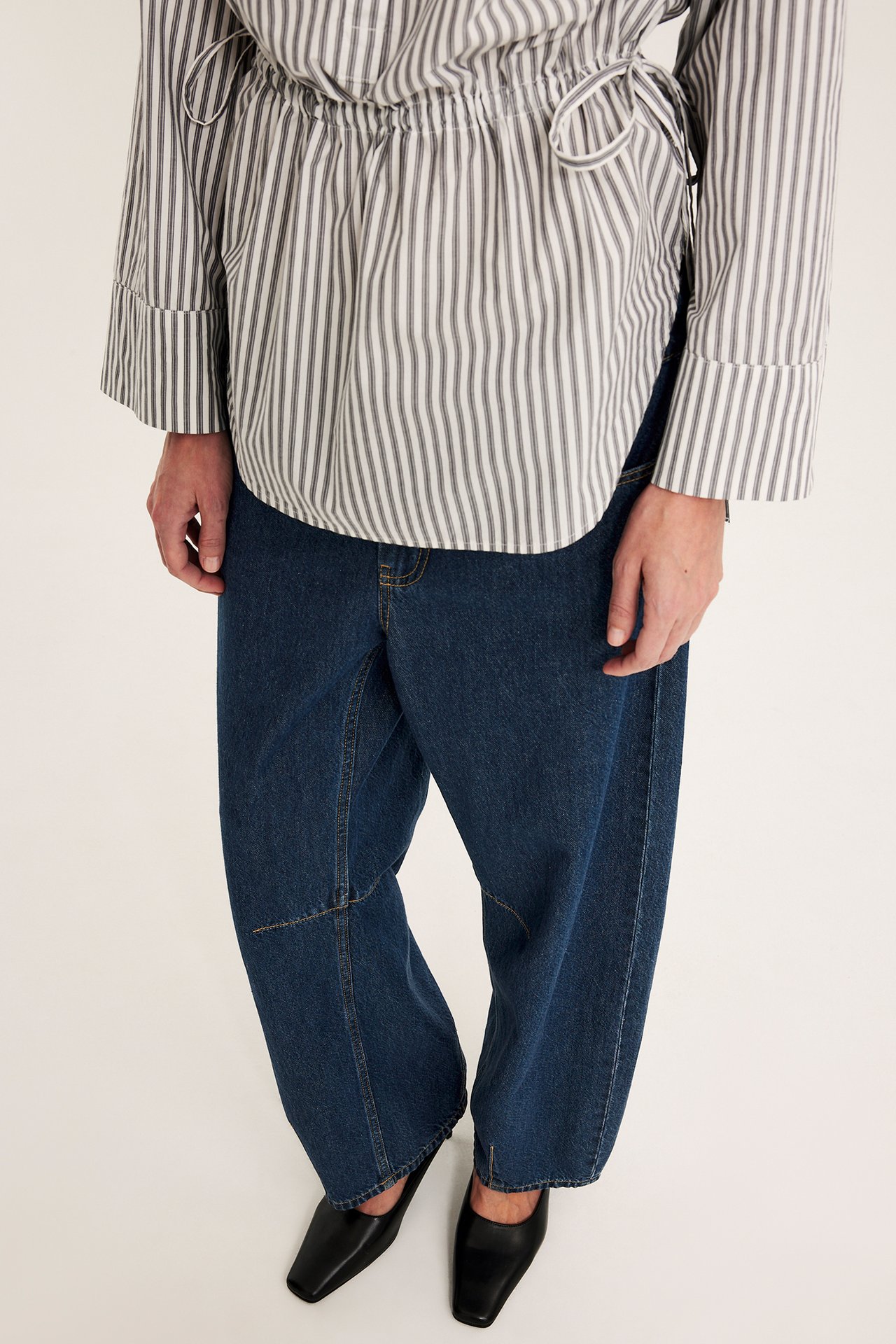 Framsida av kvinna i mörkblå barrel-fit jeans, randig blus och svarta fyrkantiga skor.