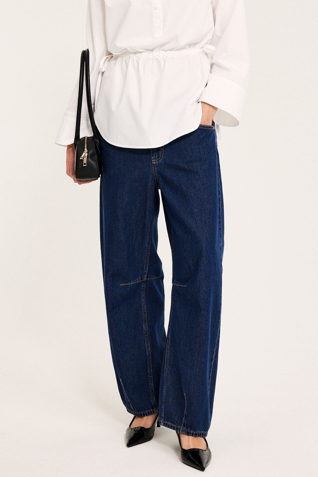 Framifrån: Kvinna i mörkblå vida barrel-jeans, vit skjorta, svart väska och svarta klackar.