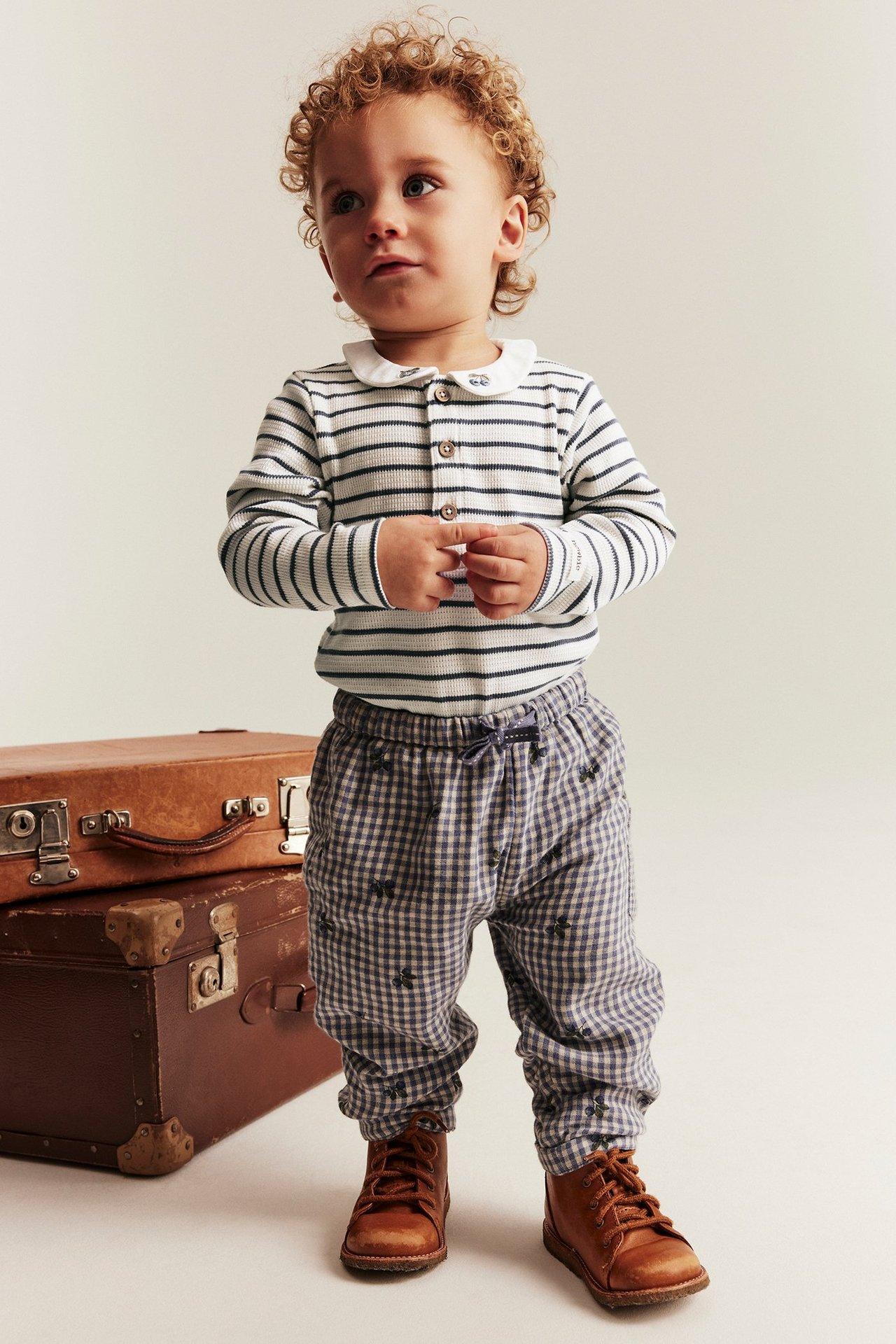 Frontbilde av baby i offwhite stripet ribbet body med brodert krage, blårutete bukser og brune boots.