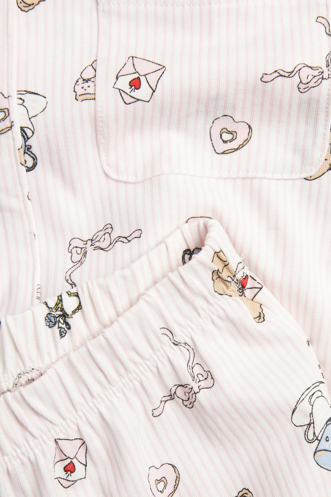 Zbliżenie: Dziecięca piżama w paski, złamana biel, z motywami kopert, kokardek i ciastek.