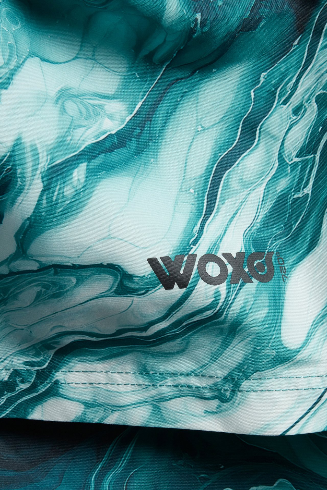 Närbild på tyg från barnens gröna och turkosa marmormönstrade badshorts med svart WOXO-logotyp.