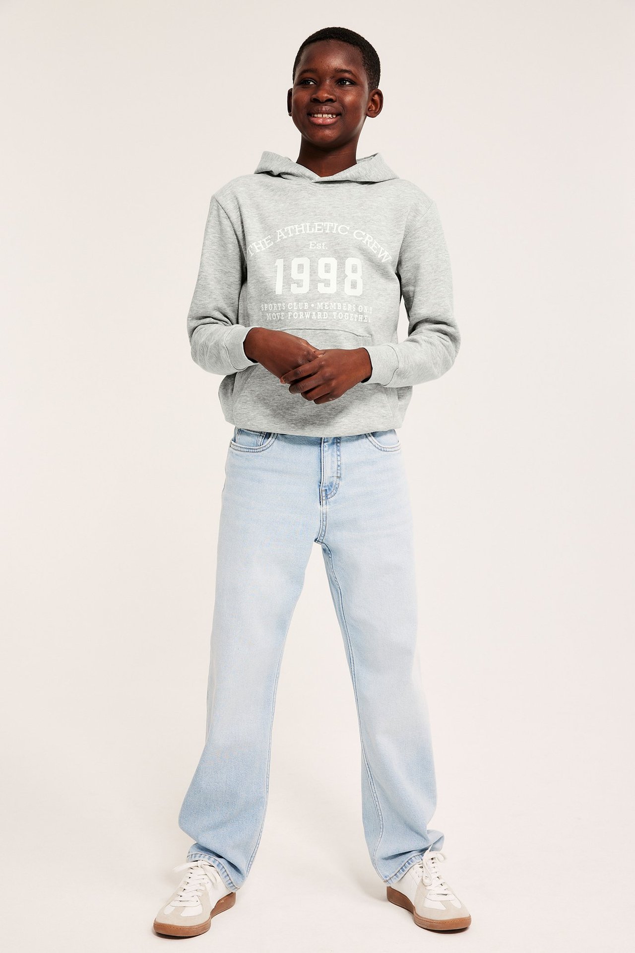 Frontvisning: Barn i gråmelert hettegenser med hvitt trykk, lyseblå jeans og hvite joggesko.