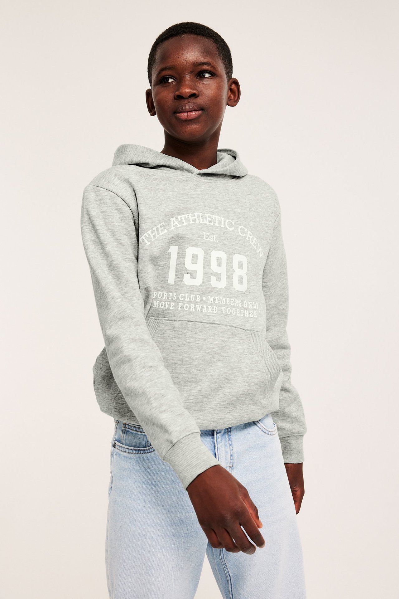 Widok z przodu: Dziecko w szarym melanżowym hoodie z białym nadrukiem i jasnoniebieskich dżinsach.