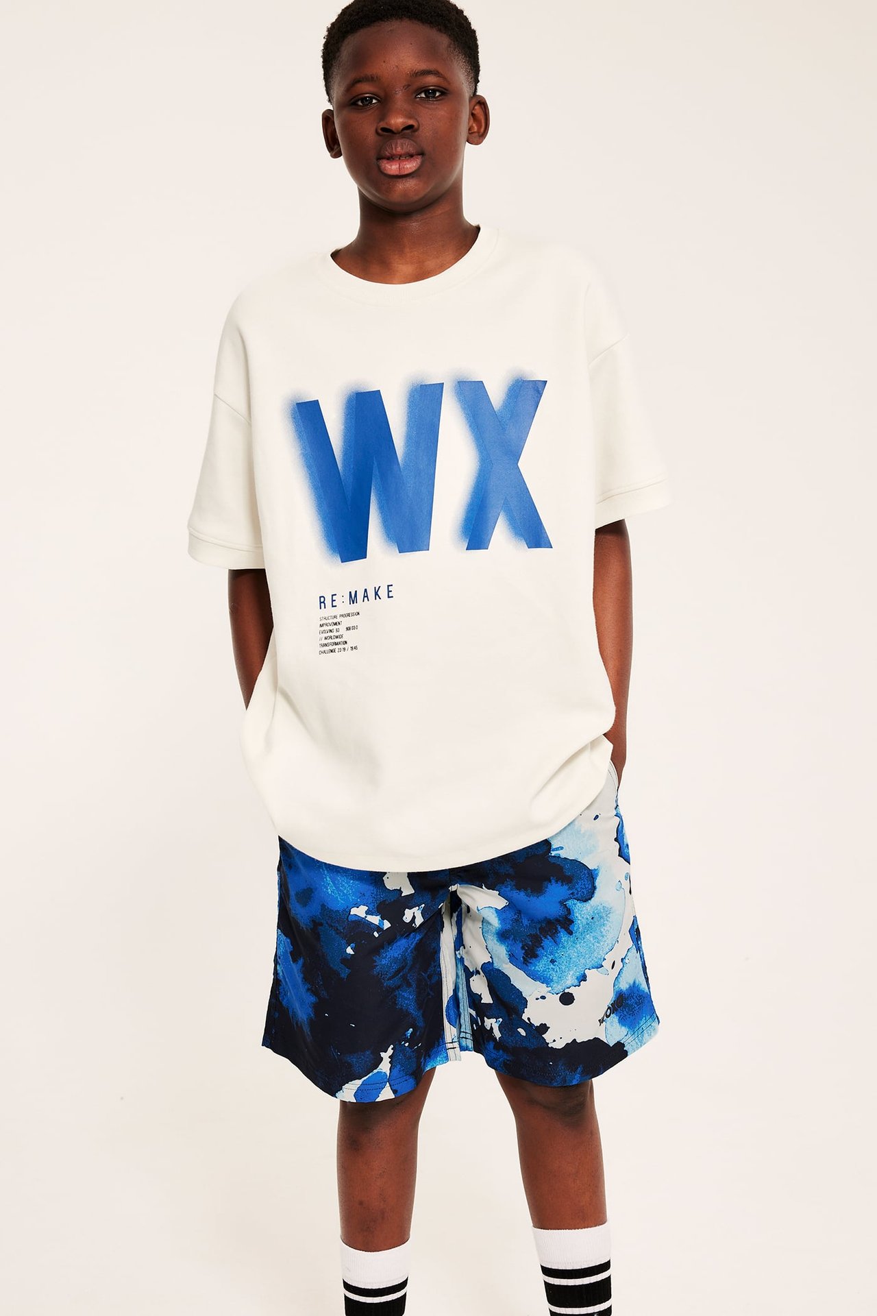 Etunäkymä lapsesta sinivalkoisissa kuviollisissa uimashortseissa ja luonnonvalkoisessa painetussa t-paidassa.