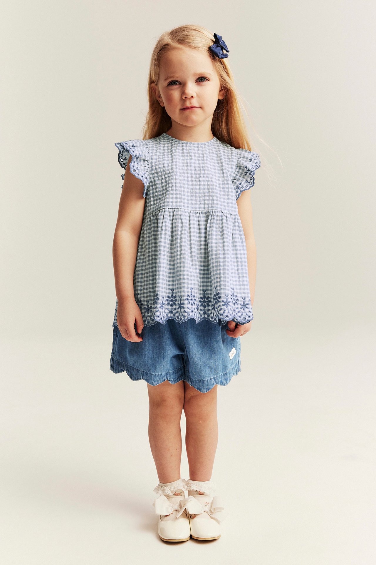 Widok z przodu: Dziecko w jasnoniebieskich szortach jeansowych z falistym brzegiem, niebieskim topie w kratkę i kremowych butach.