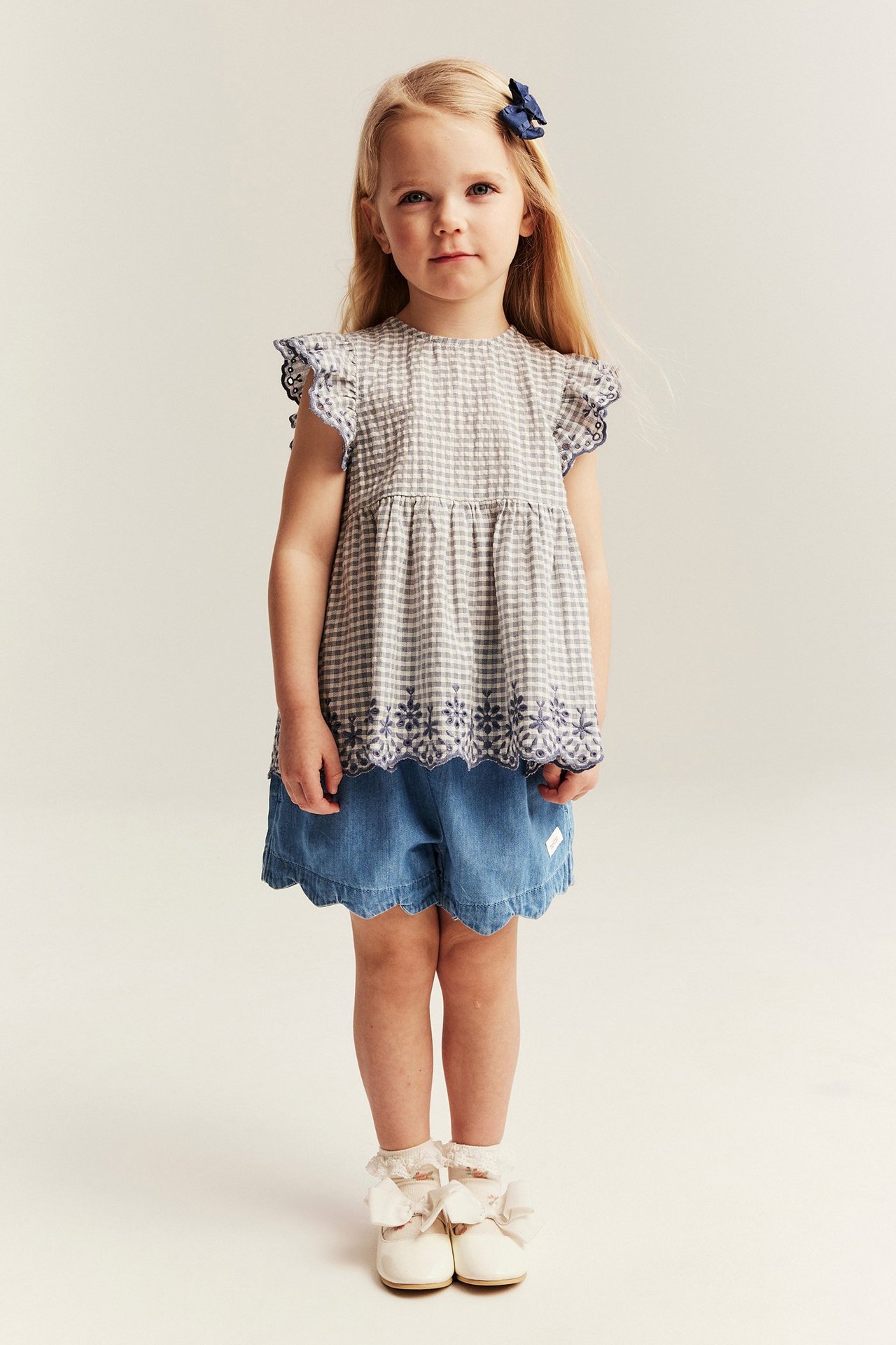 Dziecko z przodu: jasnoniebieskie szorty denimowe z falistym brzegiem, top w kratkę, niebieska kokarda, kremowe buty.