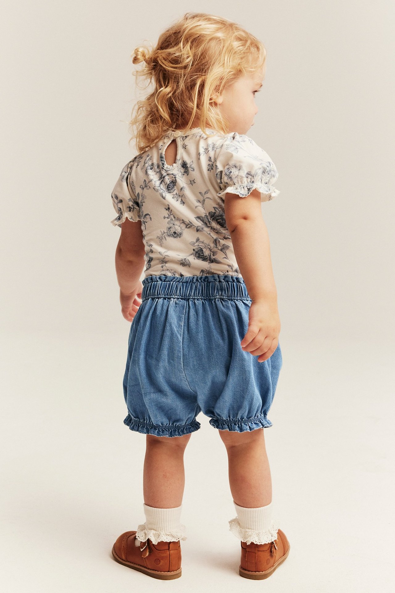 Lapsen selkäkuva vaaleansinisissä denimsortseissa röyhelöillä, valkoisessa kukkapaidassa ja ruskeissa kengissä.