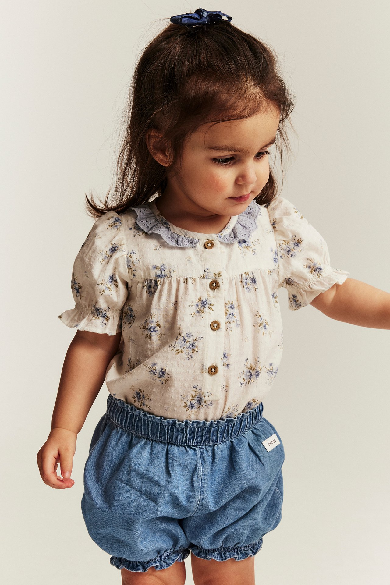 Forfra: Barn iført lyseblå denimshorts med rysjekanter, blomstret bluse og blå hårsløyfe.