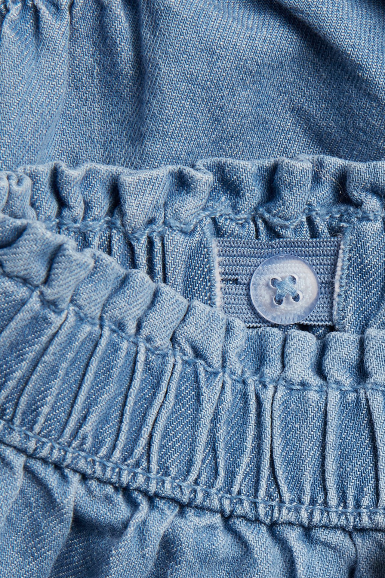 Nærbilde av lyseblå denimshorts, viser den elastiske linningen med justerbar knapp og strikk.