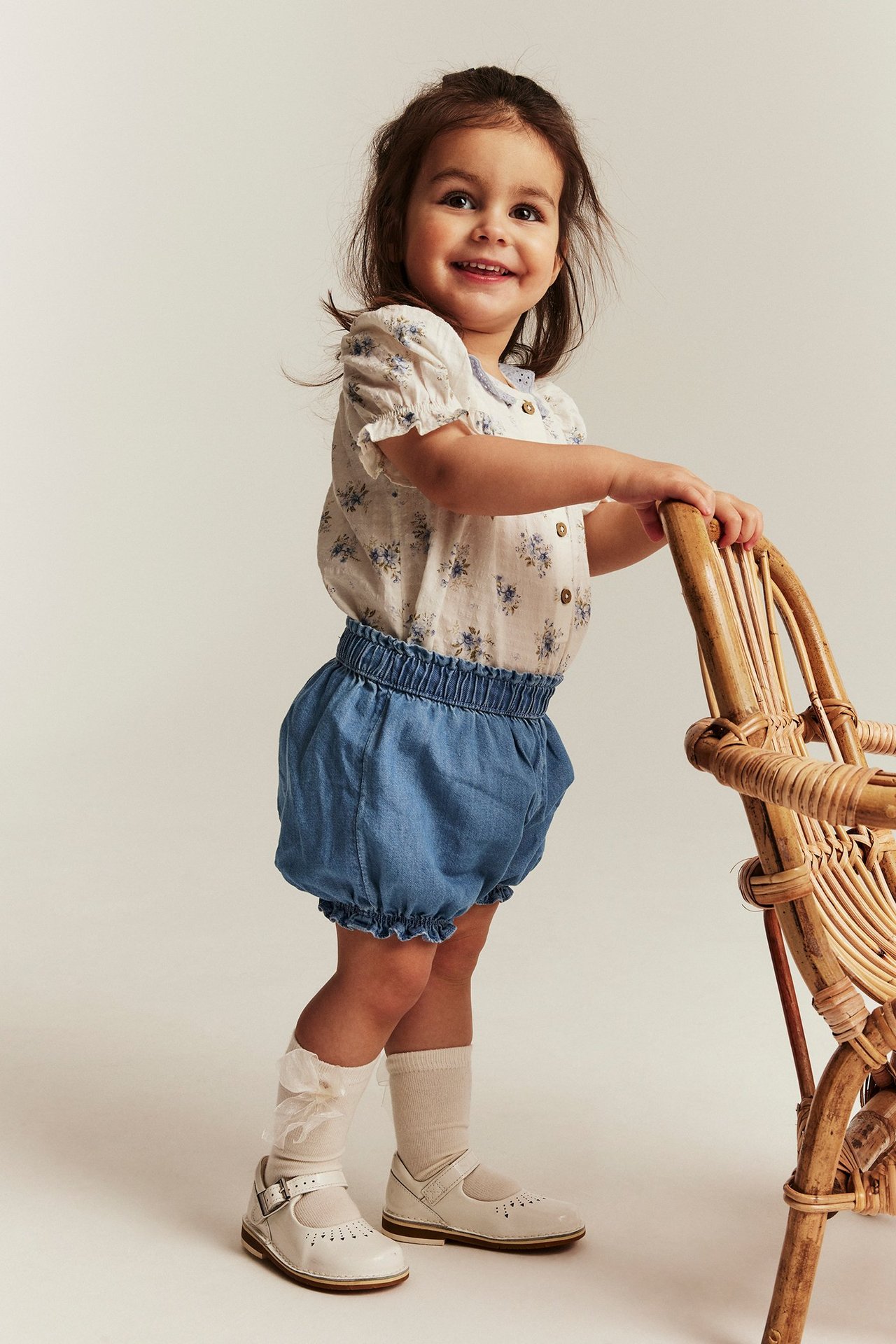 Frontbilde: Smilende barn i lyse denimshorts med strikk i midjen, blomstrete bluse, hvite sokker og sko.