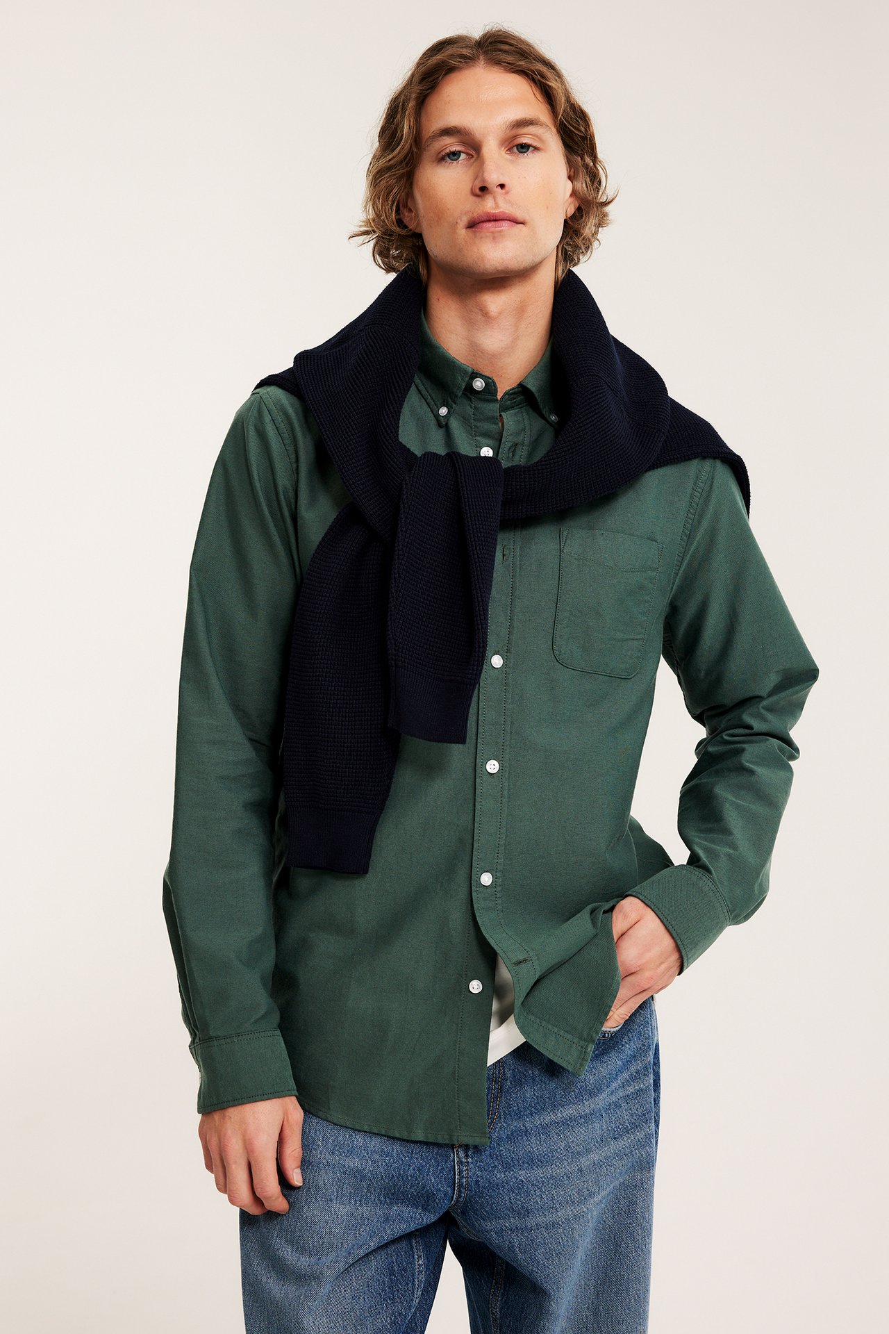 Framifrån: Man i grönmelerad oxfordskjorta, marinblå stickad tröja draperad över axlarna, och blå jeans.