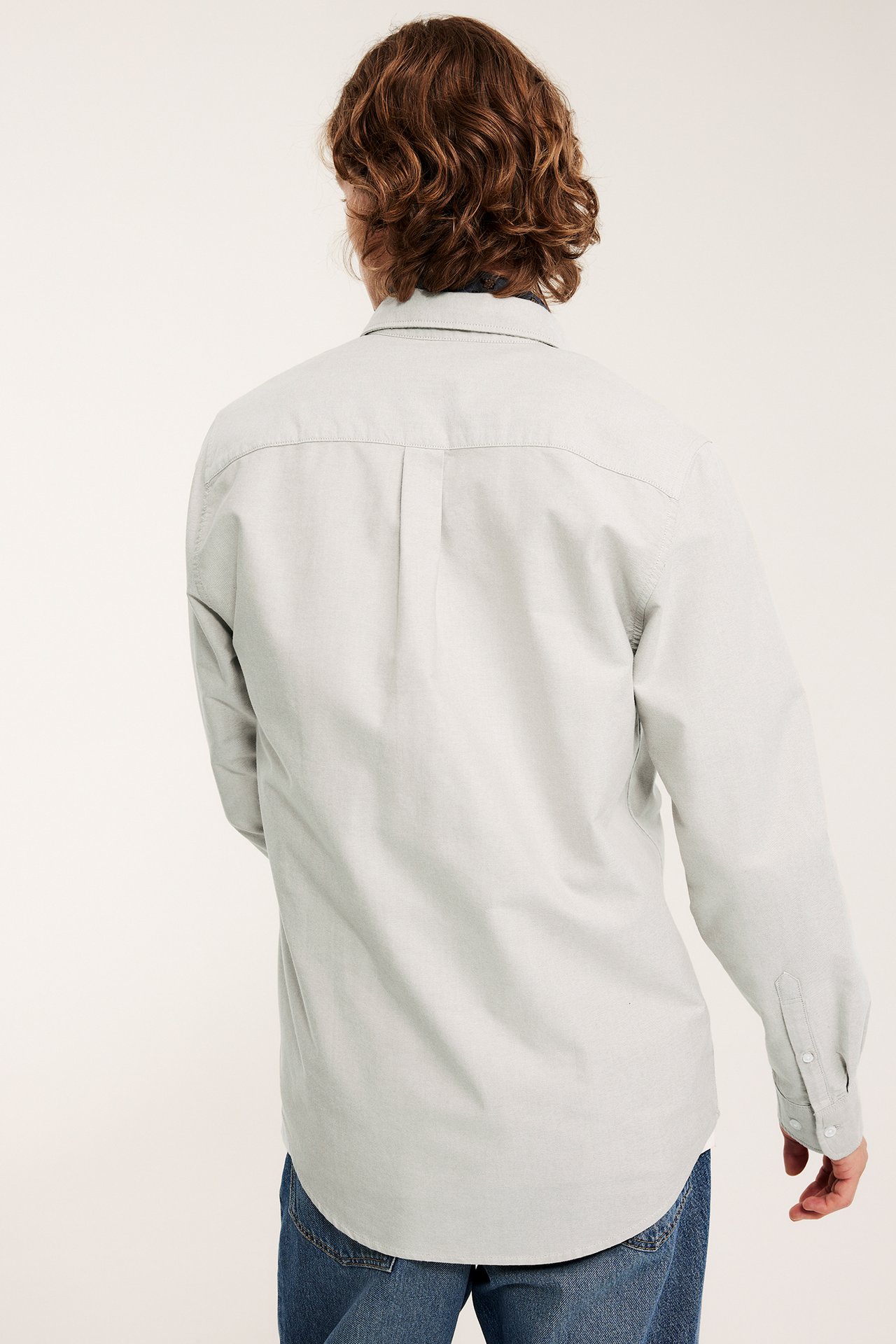 Widok z tyłu mężczyzny w jasnobeżowej melerowanej koszuli oxford regular fit i niebieskich dżinsach.