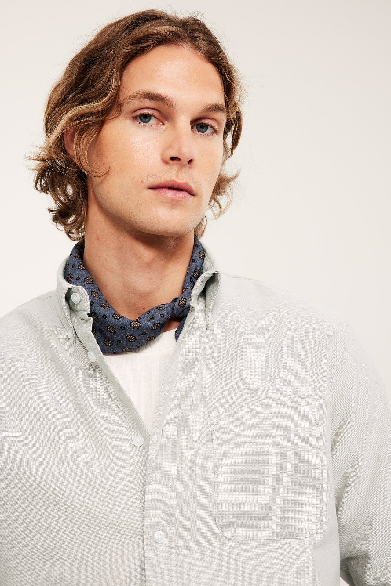 Frontvy: Man i ljusbeige melerad oxfordskjorta med button-down krage, vit t-shirt och mönstrad blå halsduk.