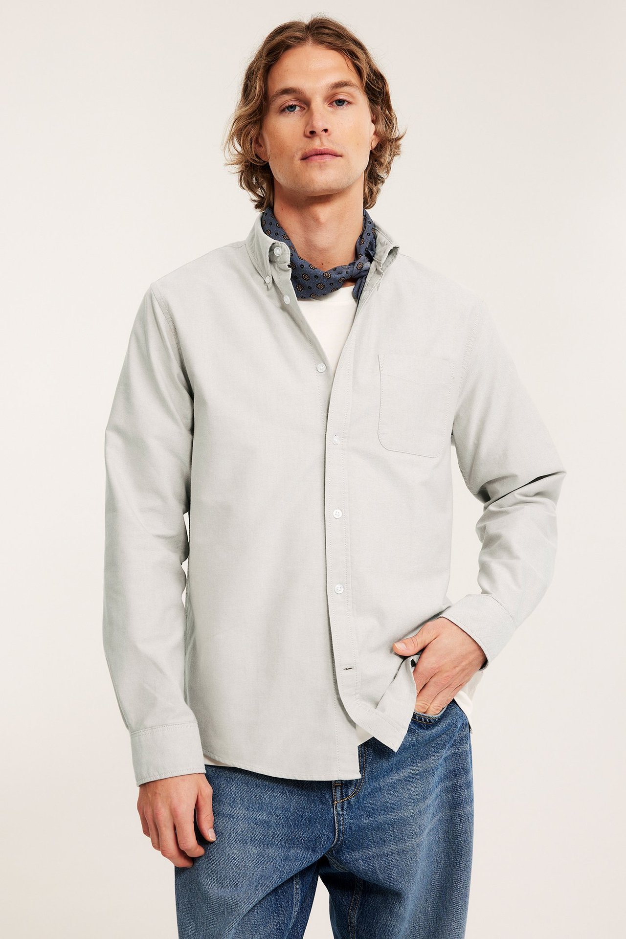 Framifrån: man i ljusbeige regular fit oxfordskjorta över vit t-shirt, blå jeans, mönstrad scarf.