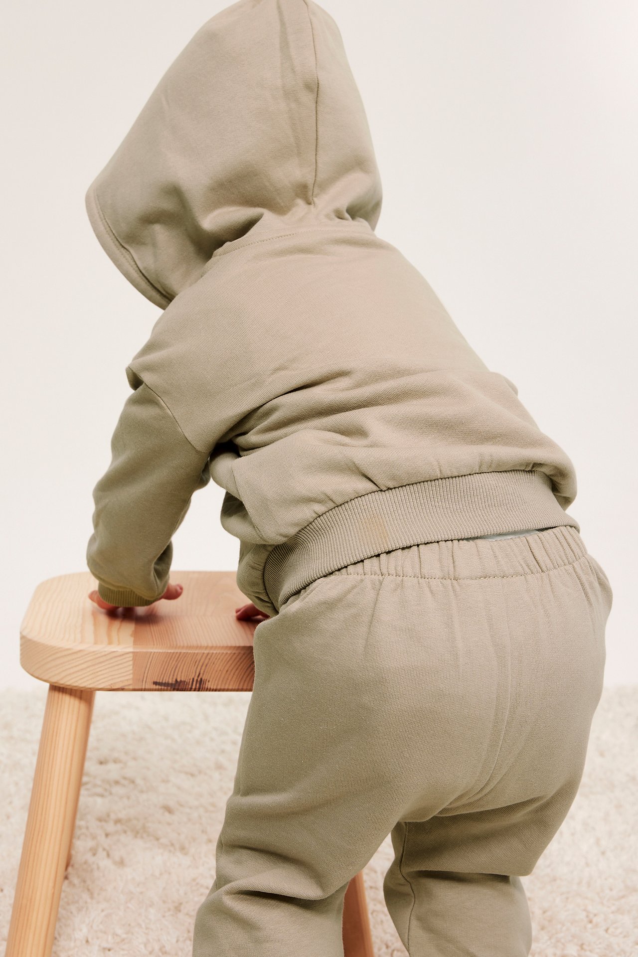 Vauva selin, beigessä puuvillahupparissa ja collegehousuissa, kiipeämässä puujakkaralle.