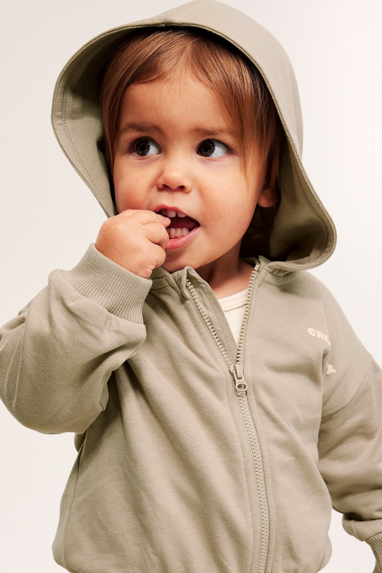 Lähikuva edestä: vauva beigessä vetoketjullisessa puuvillahupparissa, rintapainatus, vaalean paidan päällä.