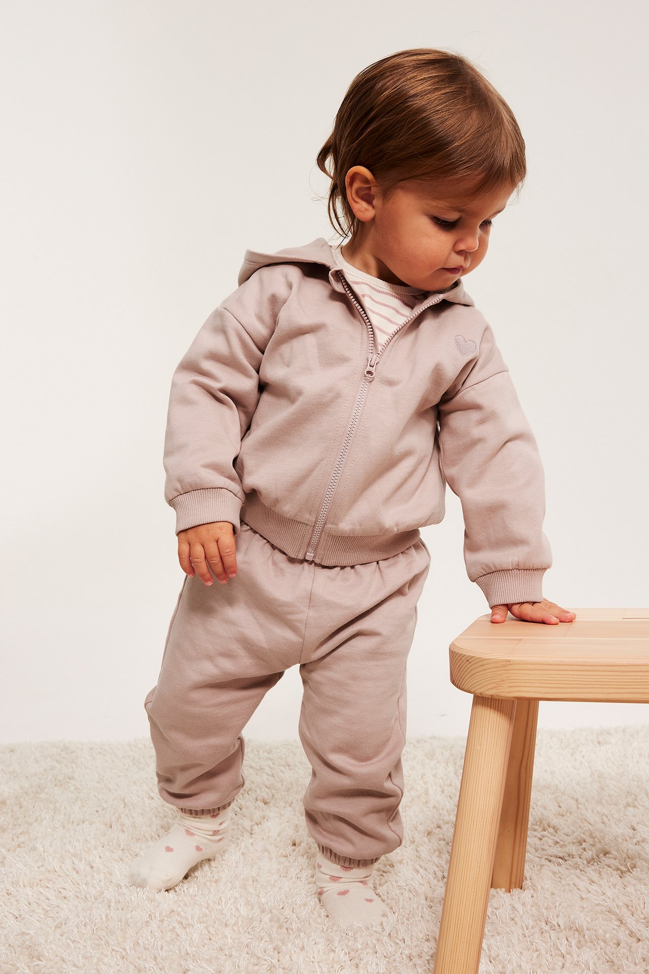 Side-forfra-visning av en baby i rosa glidelåshettgenser i bomull med brodert hjerte, matchende joggebukse og stripet genser.