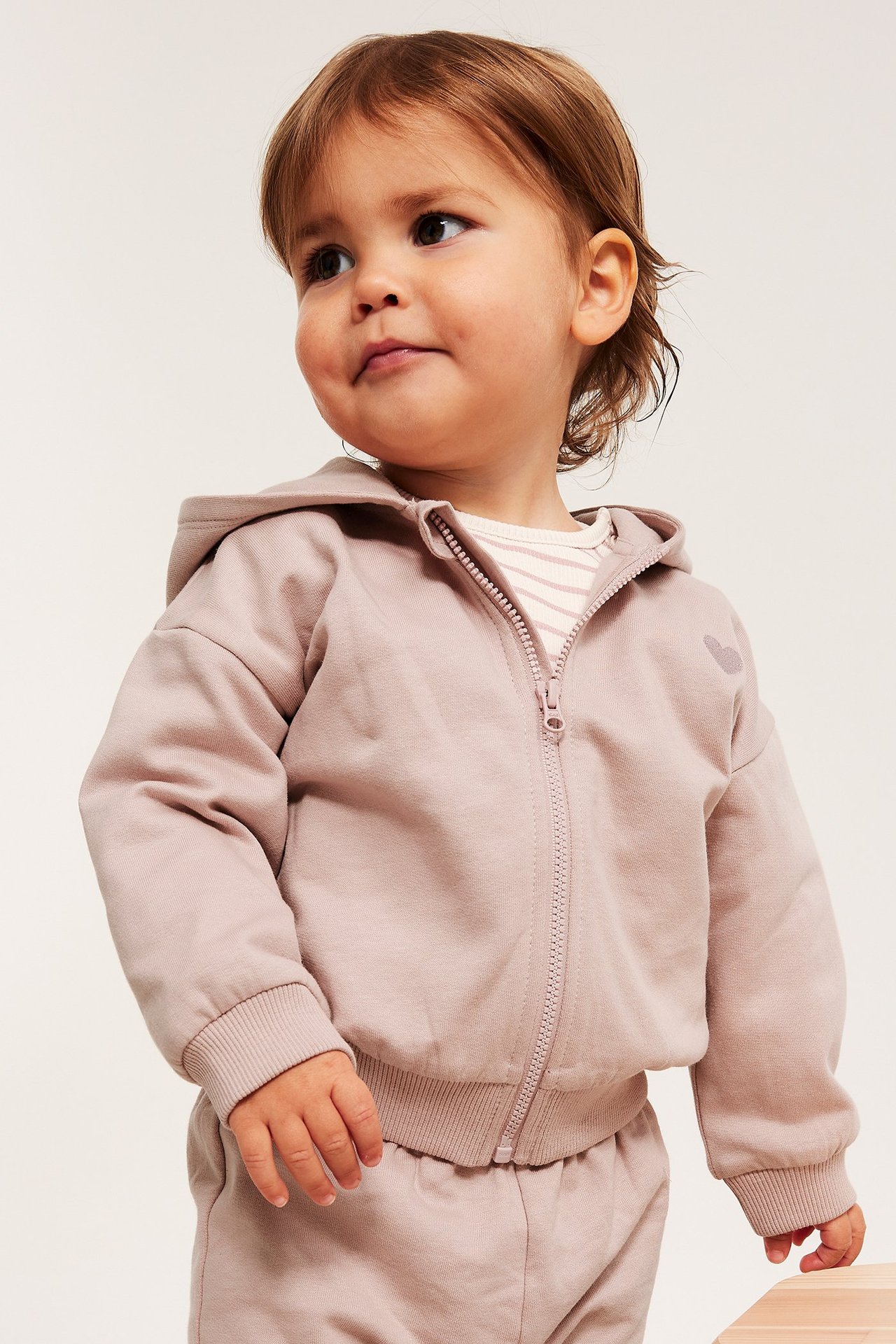 Vauva yllään vaaleanpunainen puuvillainen vetoketjuhuppari sydänbrodeerauksella, raidallisen paidan ja collegehousujen kanssa.