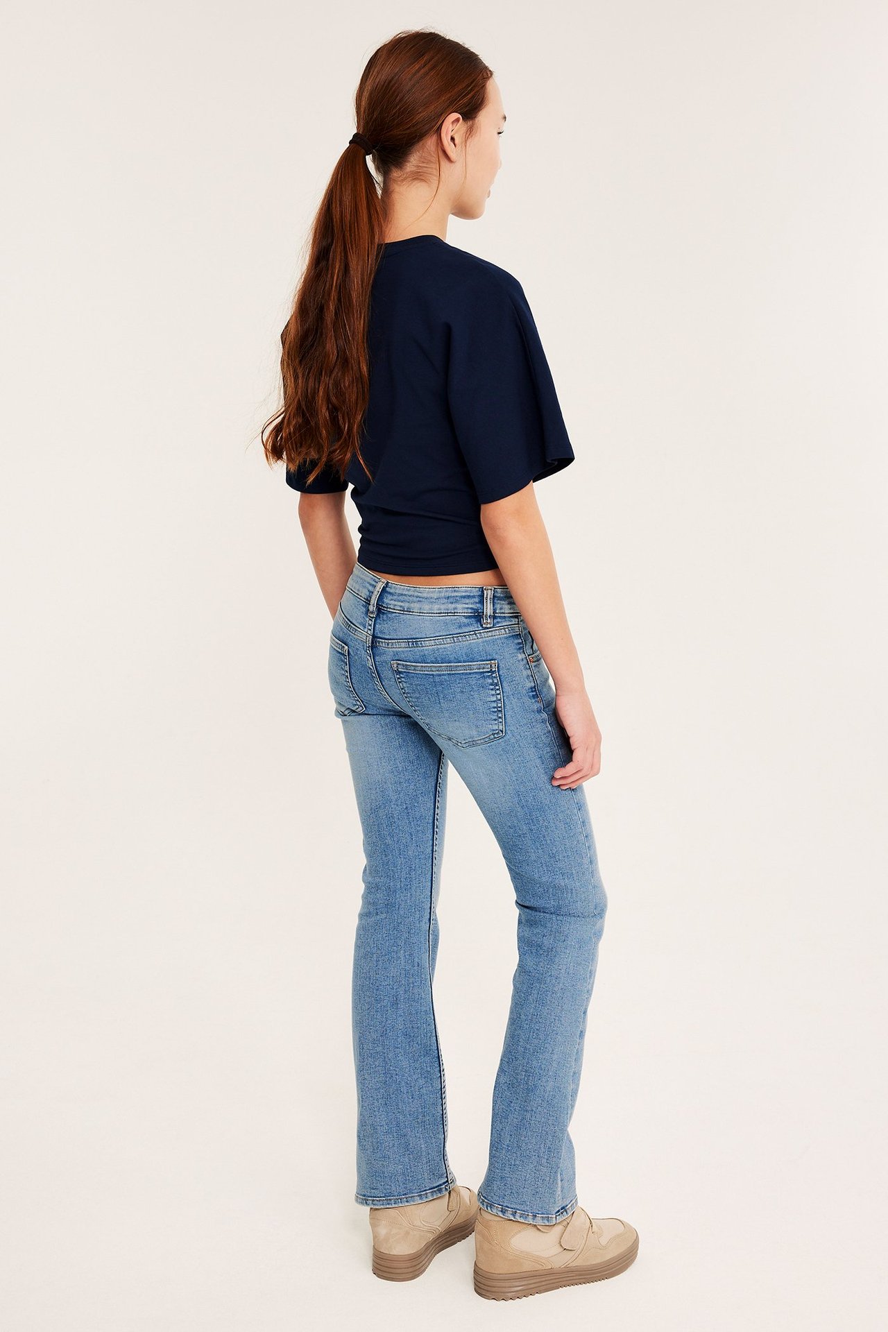 Tylny widok dziecka w jasnoniebieskich jeansach bootcut z niskim stanem, granatowym topie i beżowych botkach.