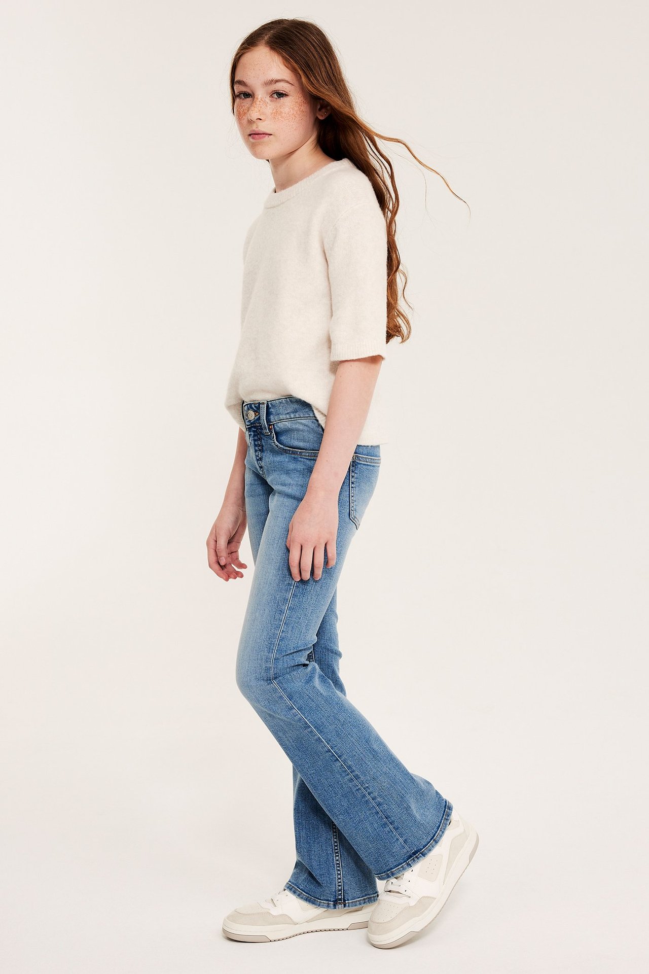 Sidovy av ett barn i blåa bootcut jeans med låg midja, en vit kortärmad tröja och vita sneakers.