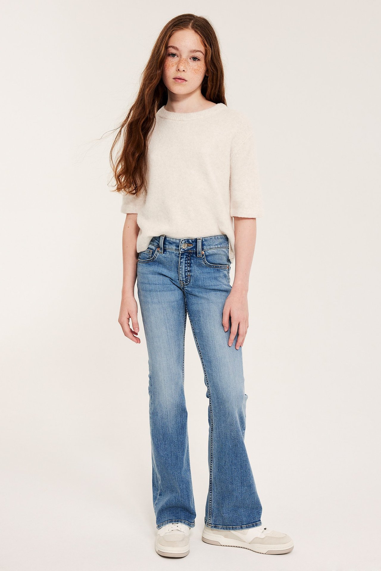 Framifrån: Barn bär ljusblå bootcut jeans med låg midja, en ljusbeige kortärmad topp och beiga sneakers.