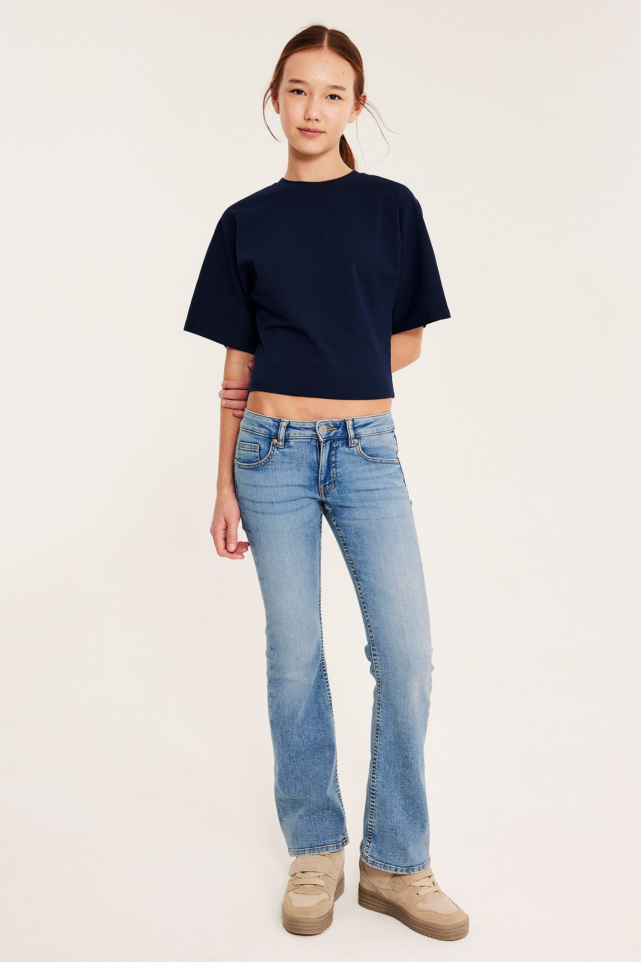 Widok z przodu: kobieta w jasnoniebieskich bootcut jeansach z niskim stanem, granatowym krótkim T-shircie i beżowych sneakersach.