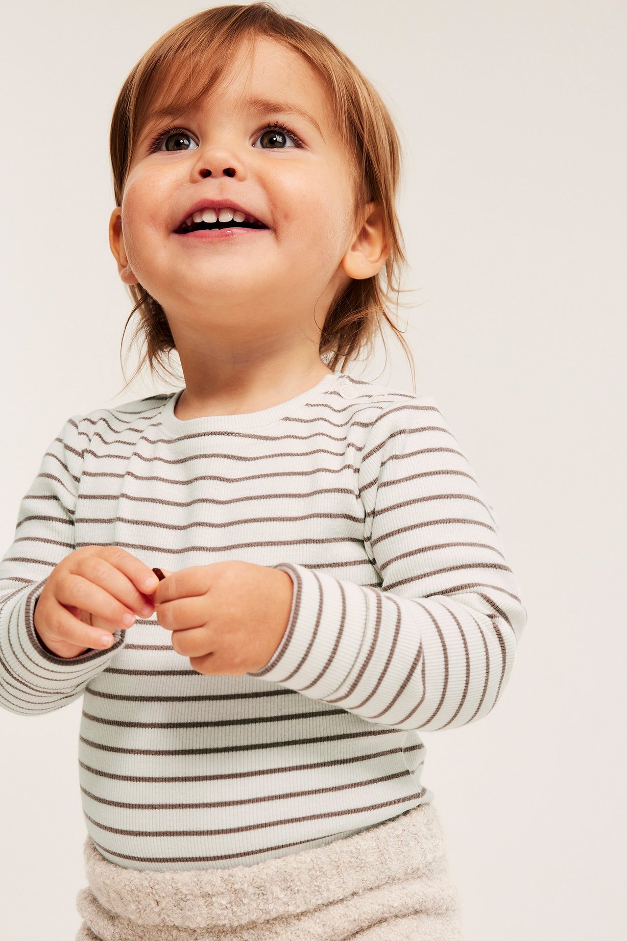 Widok z przodu: niemowlę w kremowym, prążkowanym, pasiastym body z długim rękawem i jasnobeżowych pluszowych spodenkach.