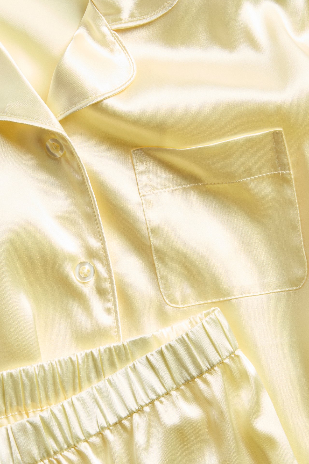 Nærbilde av lys gul satengpyjamas med knapper, krage, lomme og shorts med elastisk midje.