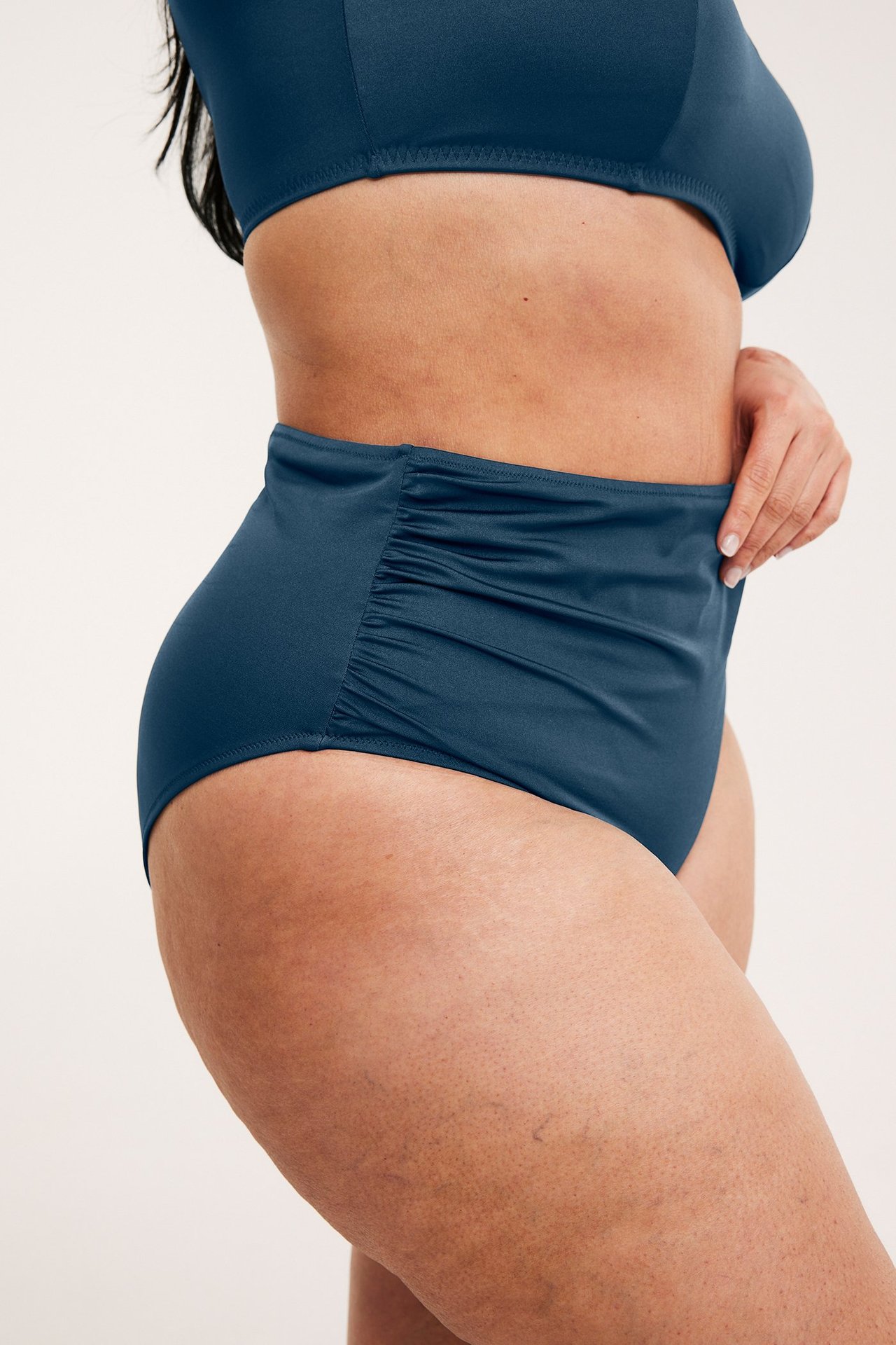 Sidebilde av en kvinne i mørkeblå høy bikinitruse med rynker i siden og en bikinitopp.