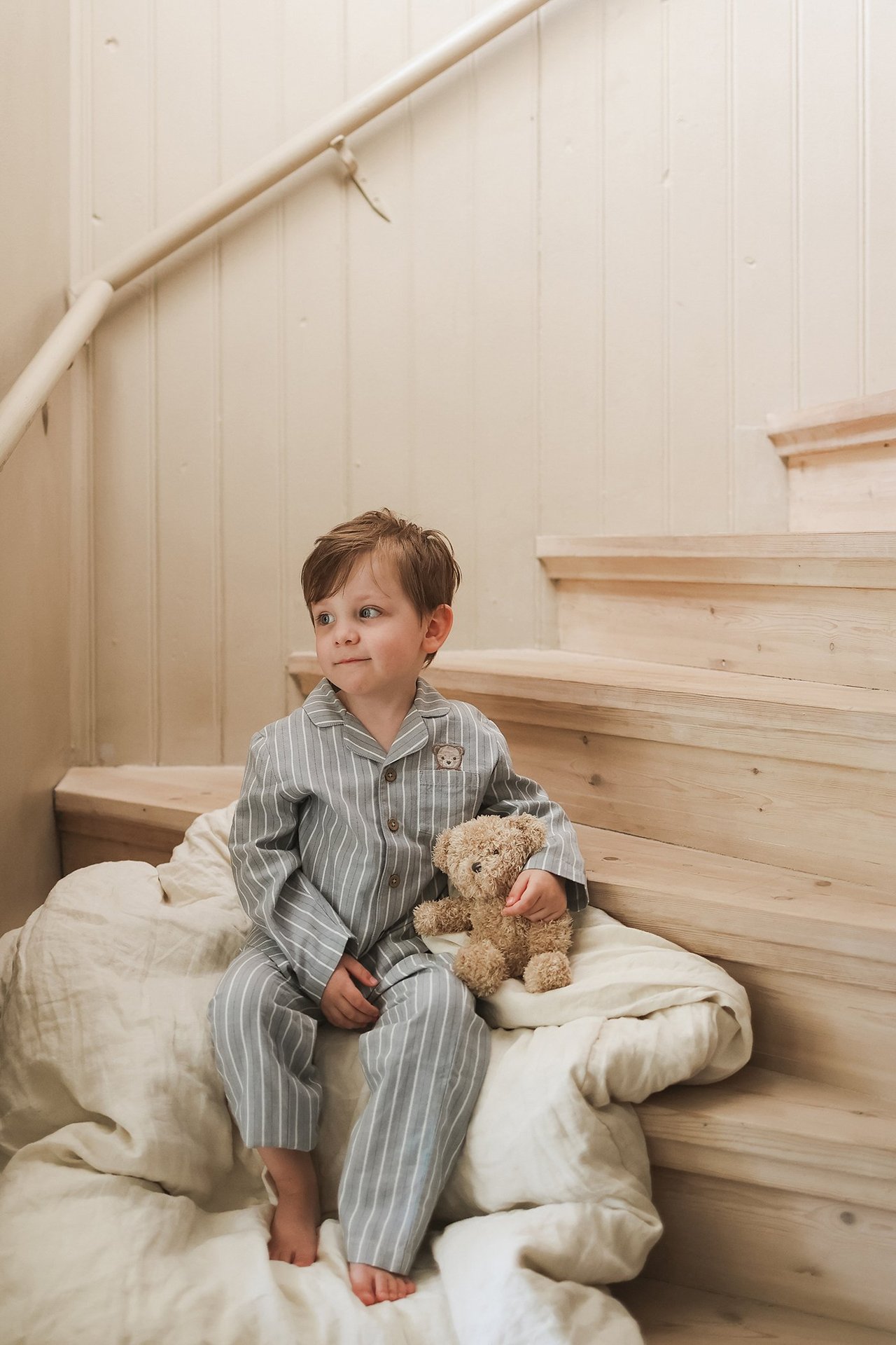 Edestä ja sivulta kuvattu lapsi vaaleansinisessä raidallisessa puuvillapyjamassa, pitelee nallea.