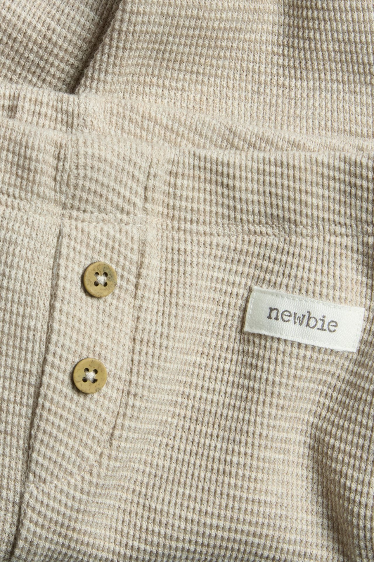 Nærbilde av beige melert vaffelstrikkede leggings, to dekorative knapper og 'newbie'-etikett.