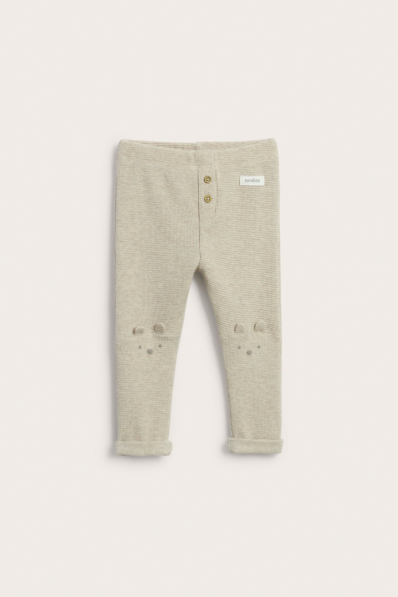 Frontalvisning: lys beige baby-vaffel-leggings med dekorative knapper, broderte kne-ører og oppbrettede ben.
