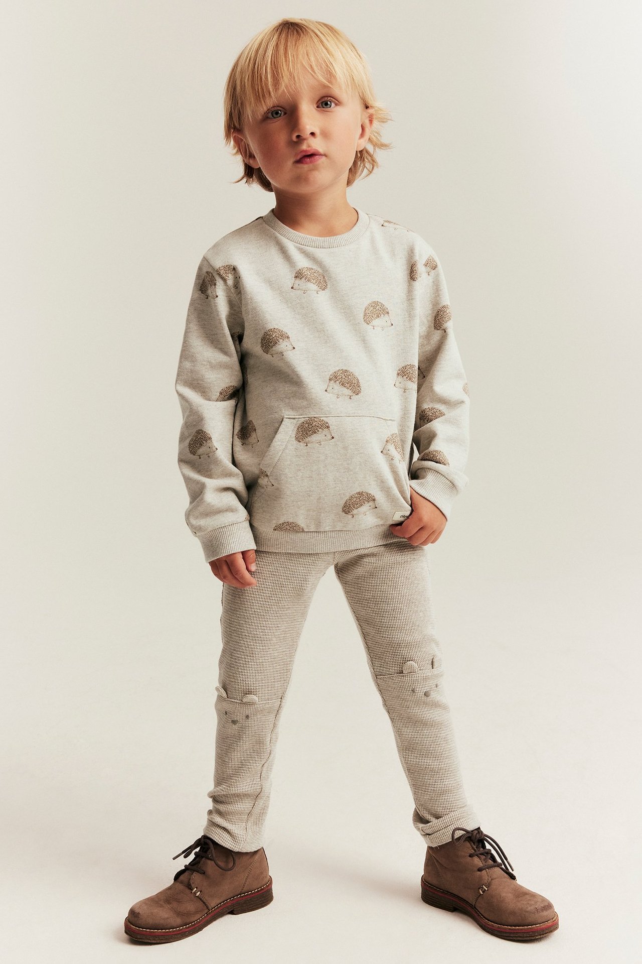 Framifrån: Barn i ljusbeige våffelleggings med öron på knäna, grå igelkottströja, bruna kängor.