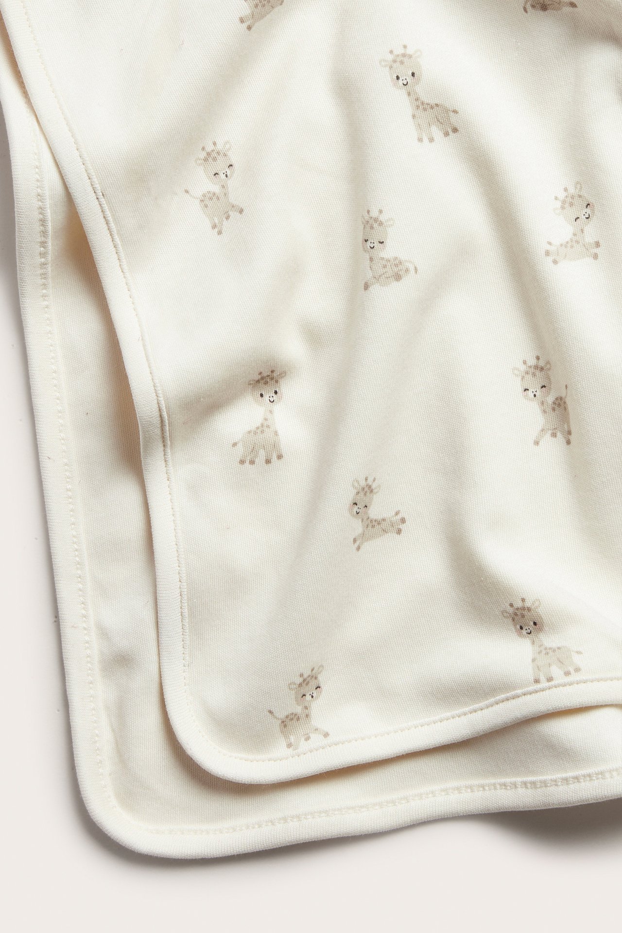 Lähikuva taitetusta vaaleanbeigestä puuvillaneuloksesta vauvan peitosta, toistuvalla kirahvikuviolla.