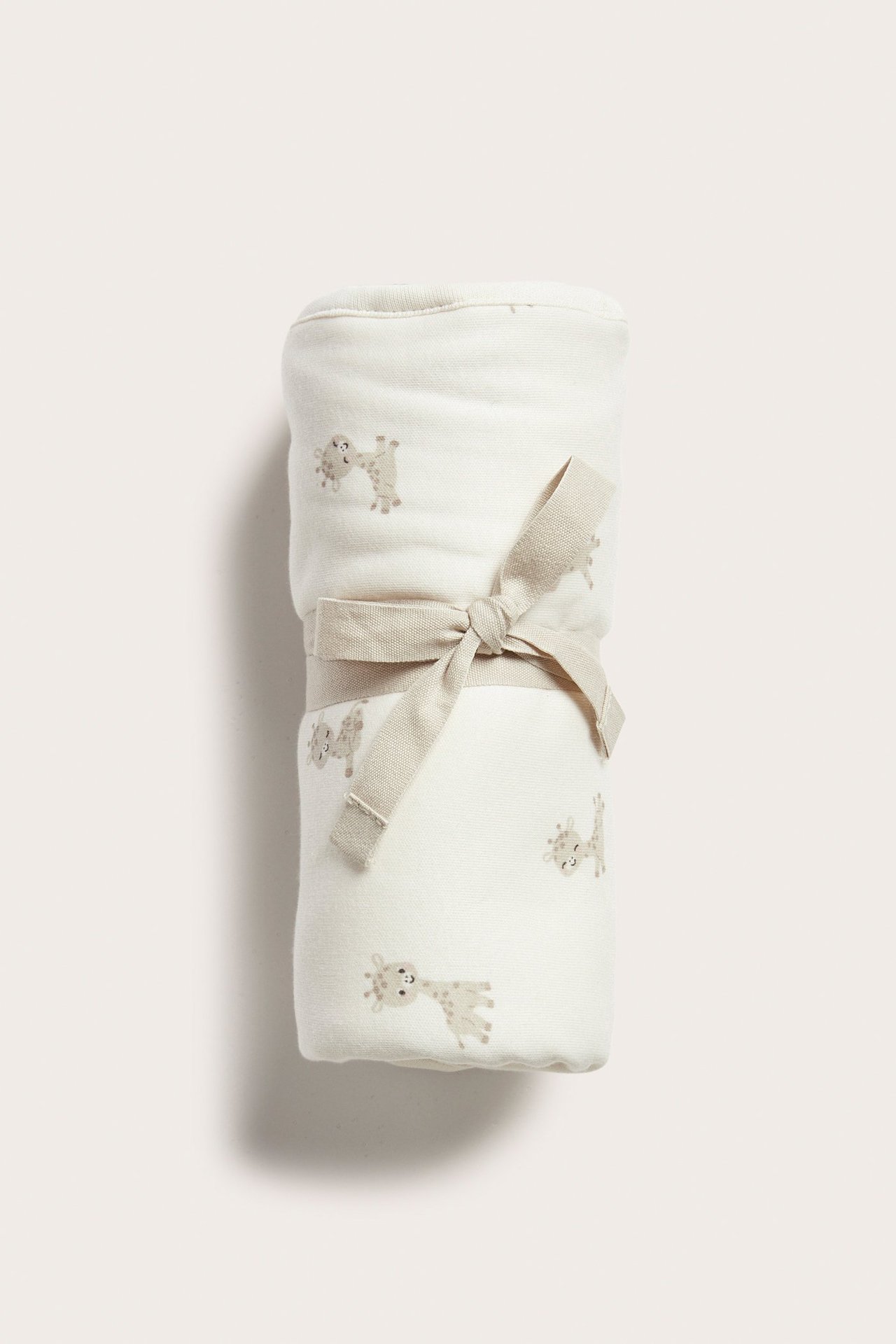 Widok z przodu: zwinięty jasnobeżowy kocyk niemowlęcy z bawełny we wzór w żyrafy, przewiązany beżową wstążką.