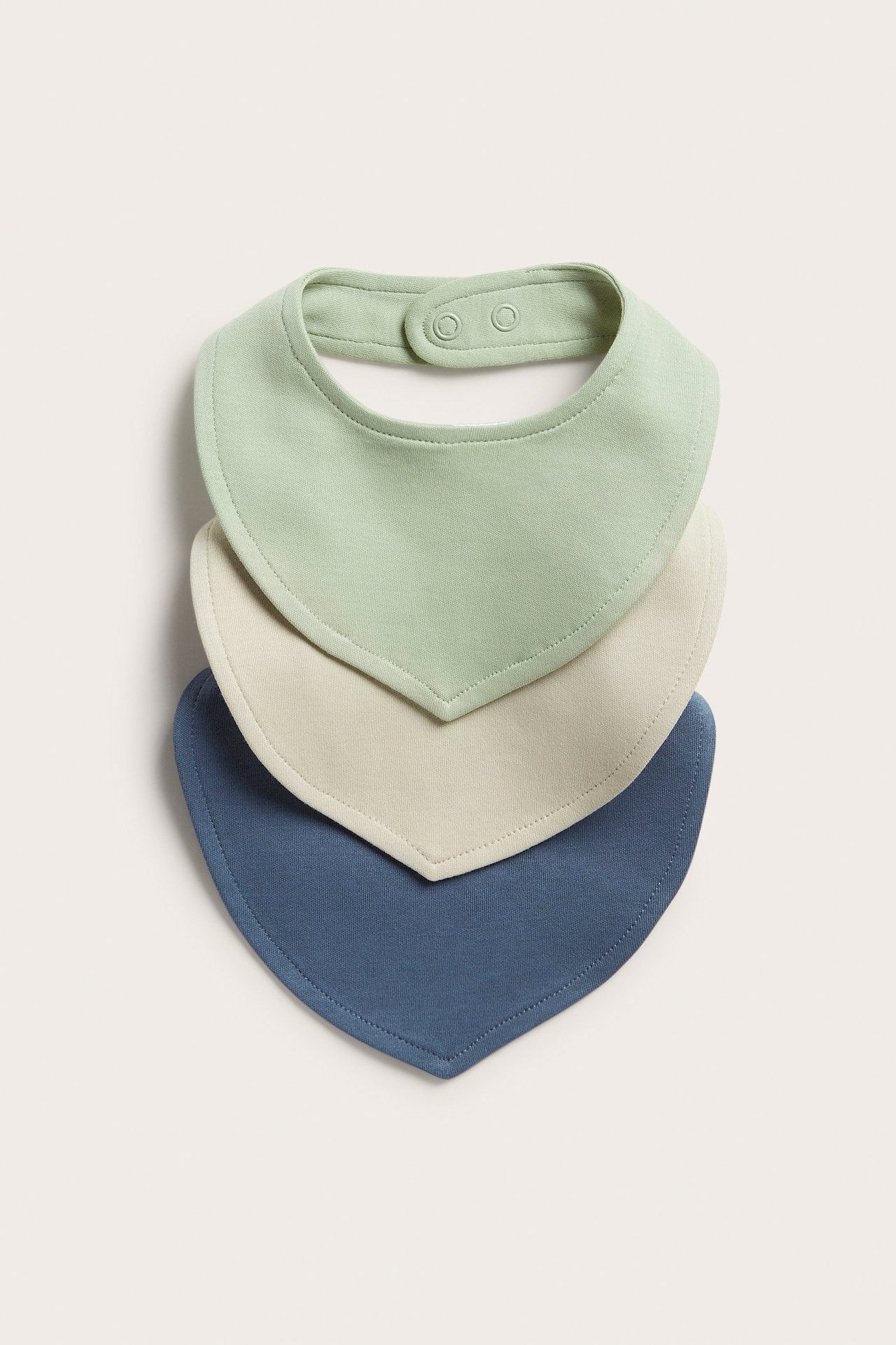 Edestä näkymä vaaleanvihreästä puuvillavauvanruokalapusta, jossa nepparit, pinottuna vaalean beigen ja sinisten lappujen päälle.