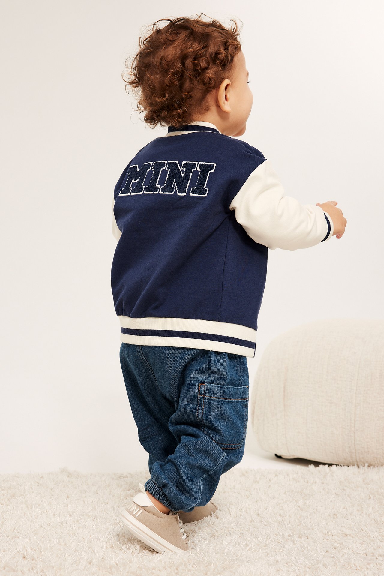 Widok od tyłu: dziecko w ciemnych dżinsach, granatowo-kremowej kurtce bejsbolówce z napisem 'MINI' i beżowych trampkach.