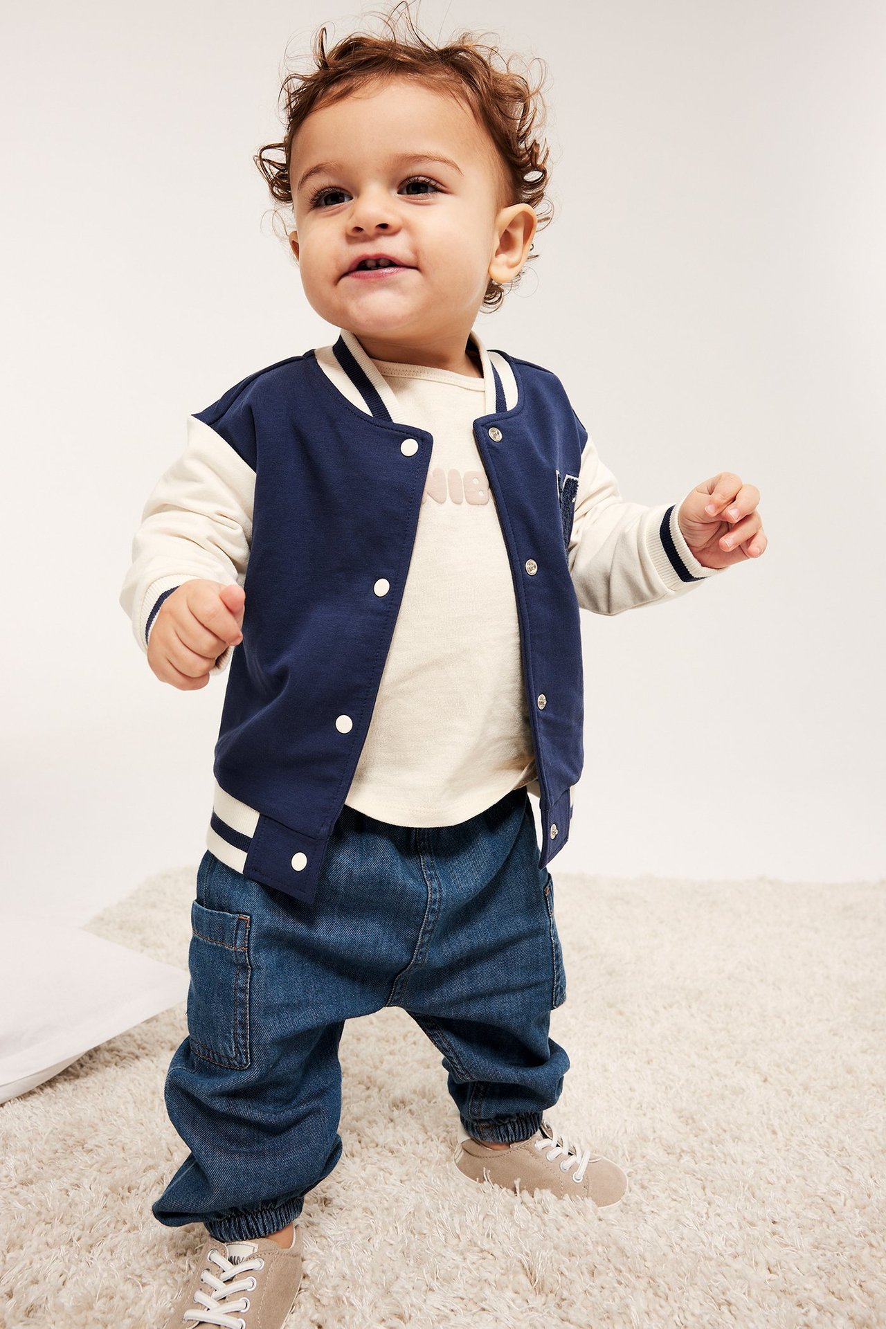 Framifrån, baby i mörkblå denimbyxor med fickor, en krämfärgad t-shirt och mörkblå collegejacka.