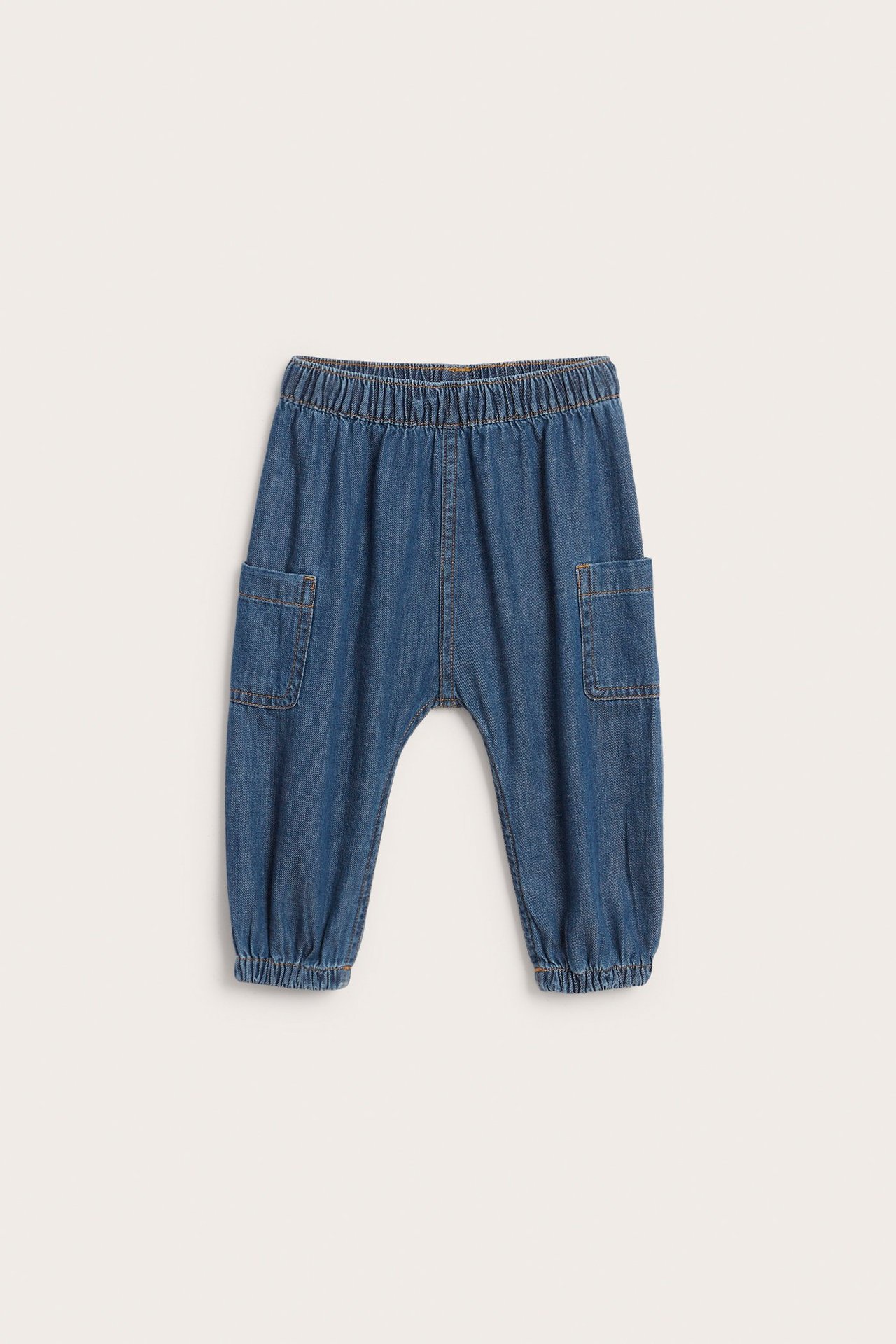 Frontvisning av baby mørkeblå denim bukser med elastisk midje, sidelommer og elastiske benåpninger.