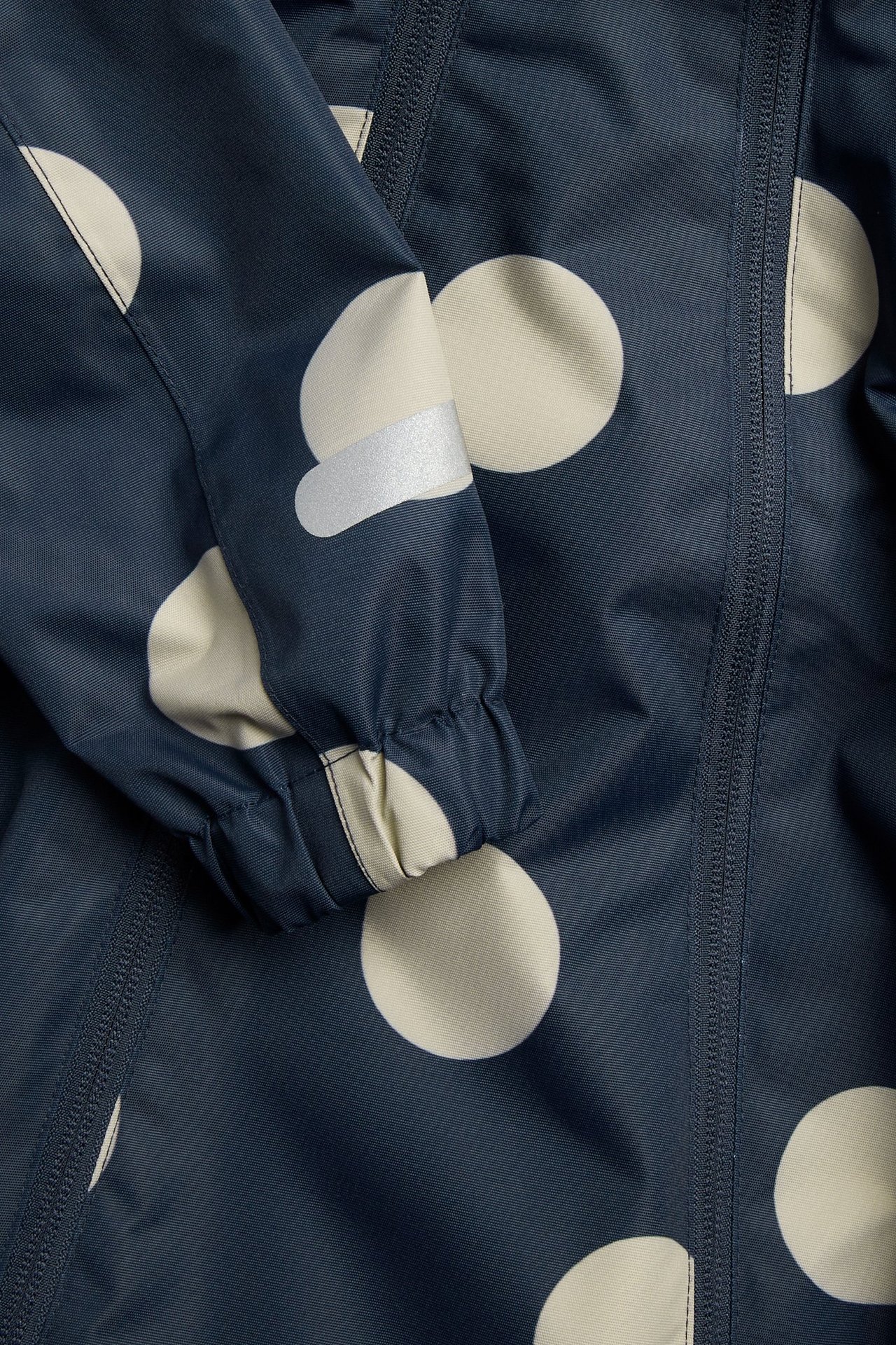 Lähikuva tummansinisestä haalarin hihasta beigenvärisillä pilkuilla, joustavalla mansetilla, heijastimella ja vetoketjulla.