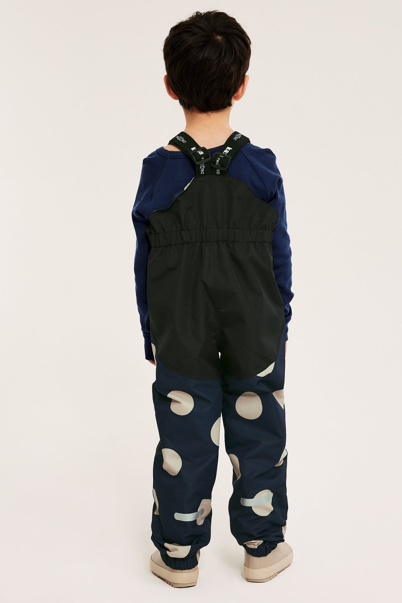 Ryggvisning av et barn i mørkeblå prikkete skallbukser med seler, blå skjorte og beige støvler.