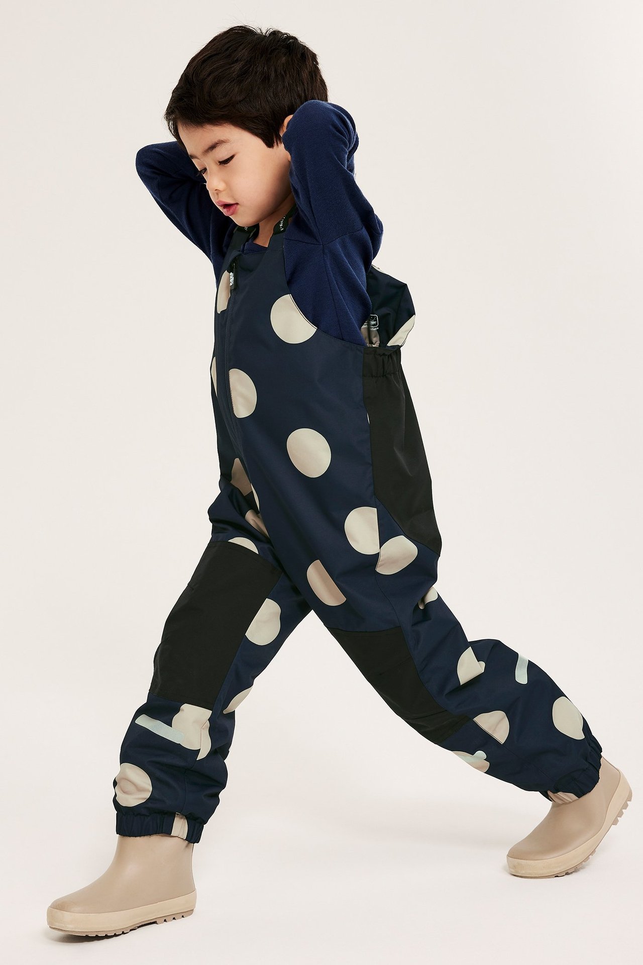 Forfra, et barn i mørkeblå prikkete skallbukse, marineblå topp og lysbeige støvler.