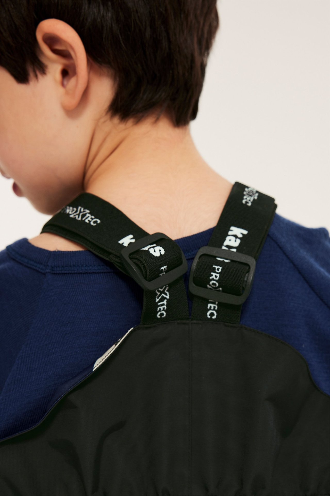 Nærbilde, bakfra av et barn i svarte Kaxs Proxtec skallbukser med justerbare merkeseler over en blå genser.