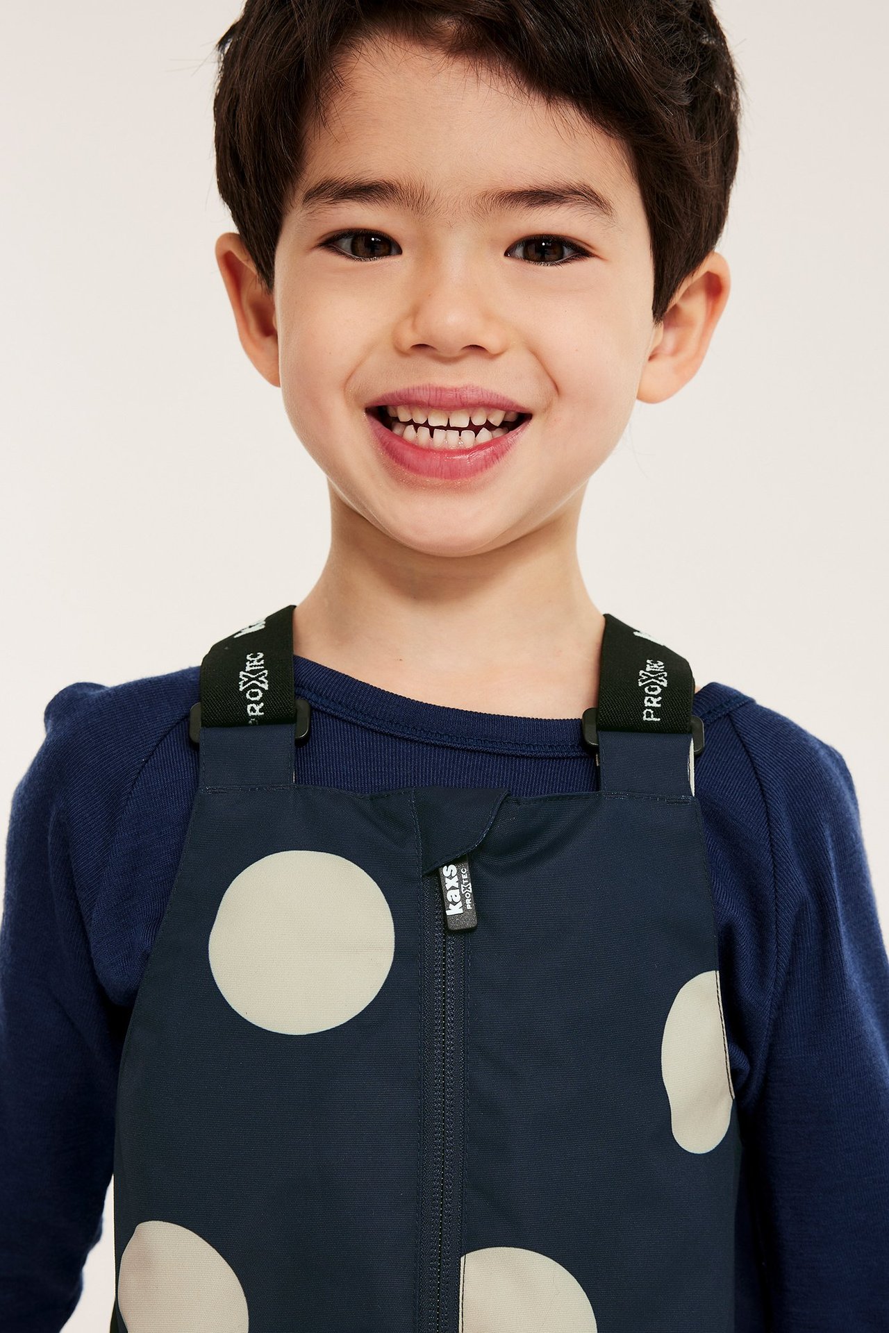 Nærbilde forfra av smilende barn i mørkeblå prikkete skallbukser med seler over mørkeblå langermet genser.