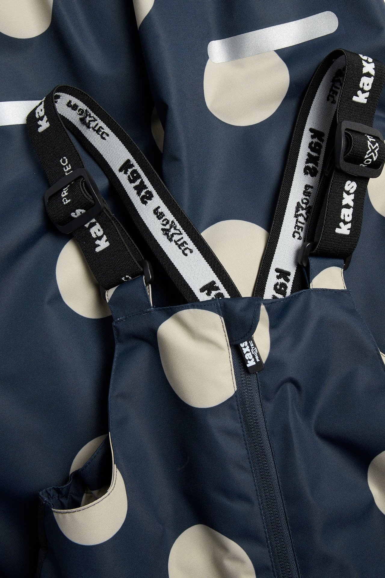 Nærbilde forfra av mørkeblå skallbukse for barn med beige prikker, justerbare svarte seler og glidelås foran.